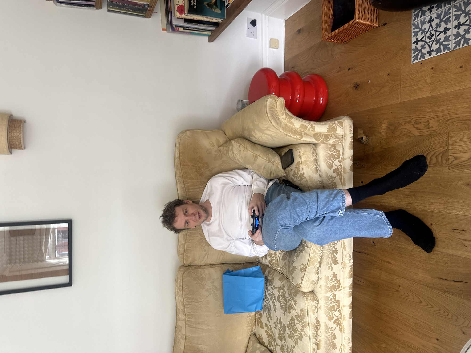 A man is lying on a patterned sofa in a room. He is wearing a long-sleeved white shirt, blue jeans, and black socks. He is holding a black game controller in his hands. Next to him on the sofa is a blue reusable shopping bag. The floor is made of wooden planks. Behind the sofa is a stack of three red, round objects and a basket. On the wall, there is a framed picture, a wall lamp, and a shelf with books. Based on the style of the room and the types of furnishings, the location is likely a residential area in Folkestone and Hythe District, United Kingdom.