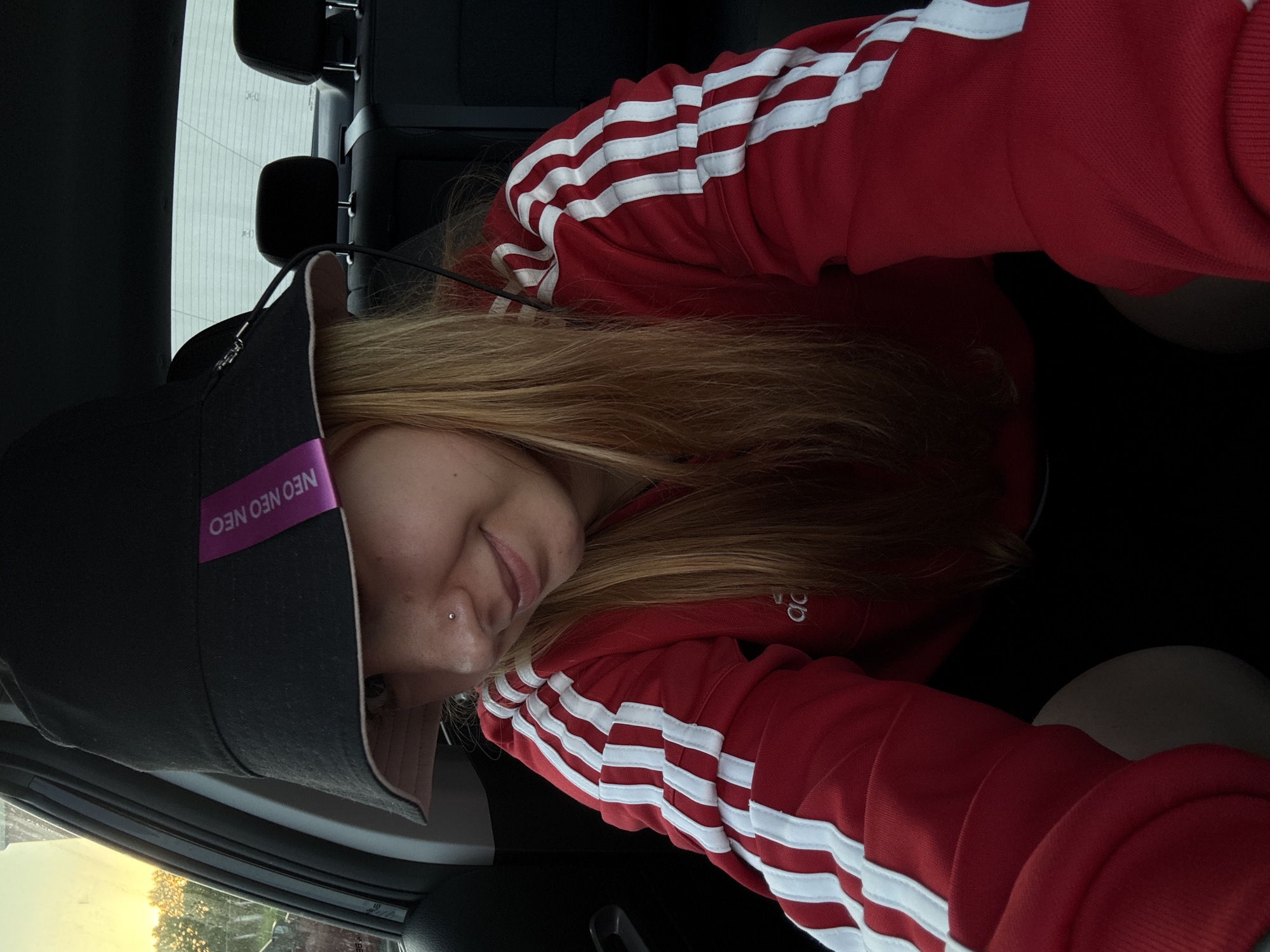 A person is sitting in a car. The individual has long, blonde hair and is wearing a black bucket hat with a pink band labeled "NEO NEO NEO". They are also wearing a red jacket with white stripes on the sleeves. The scene is taken from inside the vehicle, and the outside environment, visible through the car window, suggests an urban setting, potentially in the city of Minsk, Belarus. The person is leaning forward, looking at the camera.