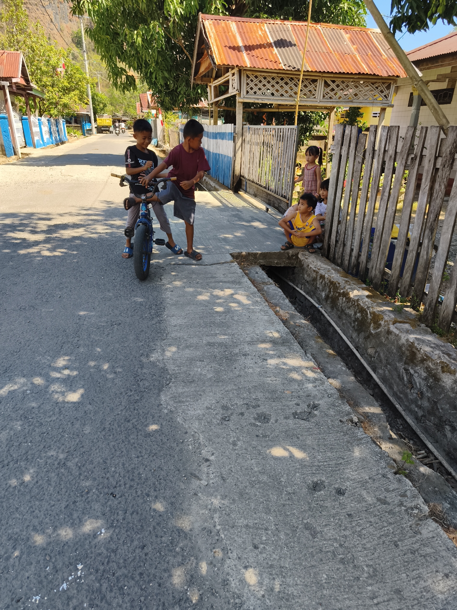 In Tonasa, Indonesia, on a sun-drenched street, a group of children are gathered. Two young boys, one in a black t-shirt and the other in a maroon shirt, stand beside a blue bicycle, appearing to be engaged in conversation or play. Further down the street, another boy in a yellow shirt crouches near a drainage ditch, looking towards the fence. Behind him, a young girl in a patterned dress stands near the fence, also observing. The street is paved, with a concrete drainage channel running alongside it. Homes with corrugated metal roofs are visible along the street, and trees provide dappled shade. In the distance, a yellow truck is parked on the road.