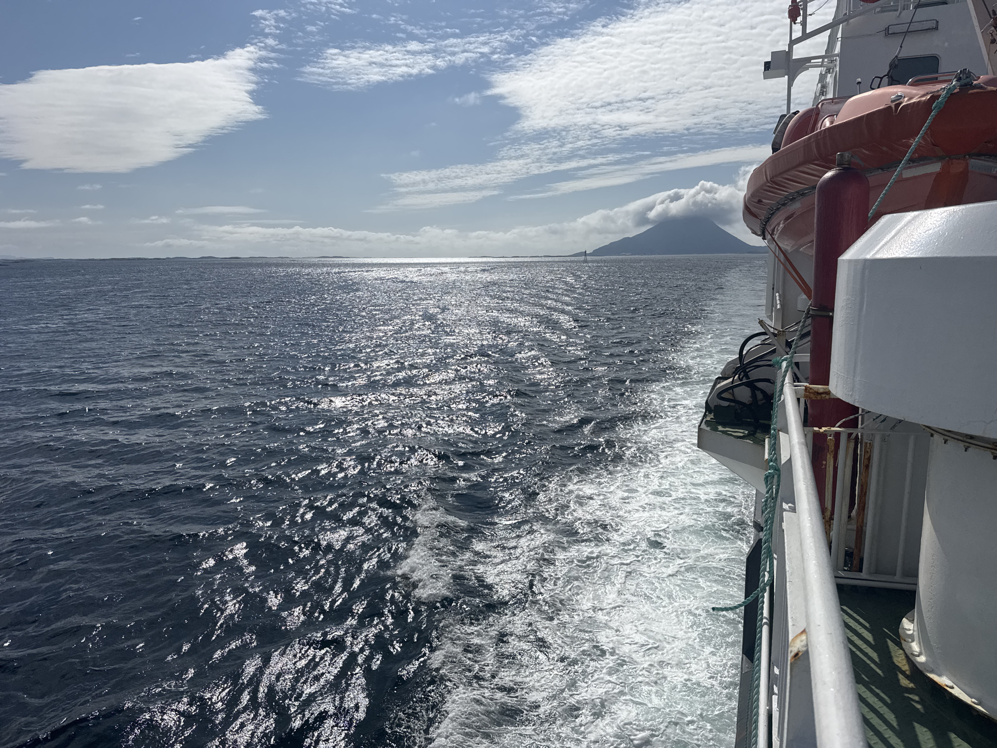 The image captures a view from the deck of a ship sailing on the ocean, likely in the vicinity of Sleneset, Norway. The water is a deep blue, disturbed by the wake of the ship, creating white frothy trails. Sunlight glints off the choppy surface of the sea, suggesting a bright, possibly windy day.
In the distance, a hazy, dark-topped mountain looms against a sky filled with scattered white clouds. The lower portions of the mountain are obscured by a layer of lighter clouds or mist. The horizon line is clearly defined, separating the sea from the distant landmass.
On the right side of the frame, the side of the ship is visible. Prominently displayed is an orange life raft, secured with ropes. The ship's structure includes white railings, white vertical cylinders, and other white structural elements. A green rope hangs down the side of the ship. The overall impression is of maritime travel in a scenic, possibly remote, coastal environment.