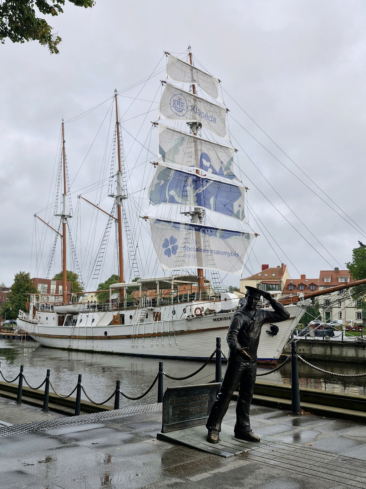 The image depicts the Meridian sailing ship docked in the city of Klaipėda, Lithuania. In the foreground, a bronze statue of a sailor stands on a wooden platform, looking out towards the water. The sailor statue is dressed in uniform with a cap and is positioned as if shielding his eyes to see something in the distance. A dark metal plaque is visible at the base of the statue.
Behind the statue, the Meridian, a white sailing ship with multiple masts, is docked along a quay. The ship's sails are partially unfurled and display blue and white patterns, some with text. The ship has a traditional design with a white hull and wooden accents.
The environment is a waterfront setting. The quay in the foreground is paved with grey stone and exhibits wet patches, suggesting recent rain. A black metal fence with ropes separates the quay from the water. The water reflects the overcast sky and the ship. In the background, buildings with red-tiled roofs and trees are visible. The sky is cloudy and grey, indicating an overcast day. The overall scene conveys a maritime atmosphere within an urban environment.