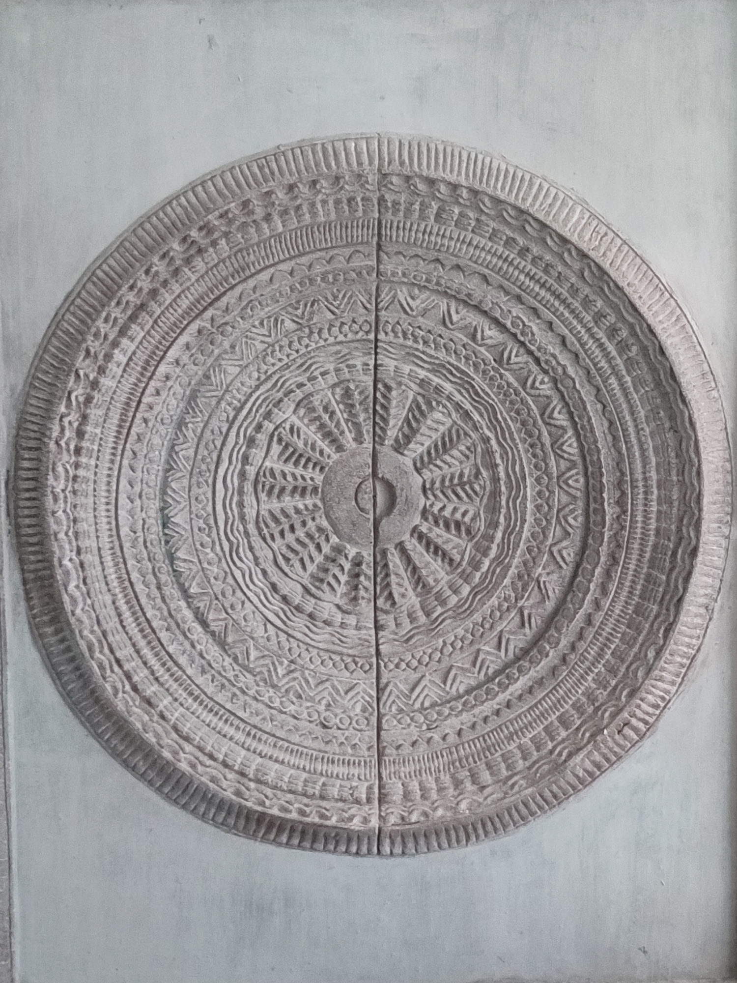 The image depicts a large, circular stone carving affixed to a wall, possibly a door or architectural element. The carving appears to be comprised of two halves, joined vertically down the center. It features intricate concentric patterns, including geometric designs such as triangles and zigzags, as well as radial elements emanating from a central hub. The overall impression is of an ancient or tribal decorative motif.
While the prompt mentions Pattaya City, Thailand, the image itself does not provide enough information to independently verify its location. Therefore, the description is limited to the visible elements in the photograph.