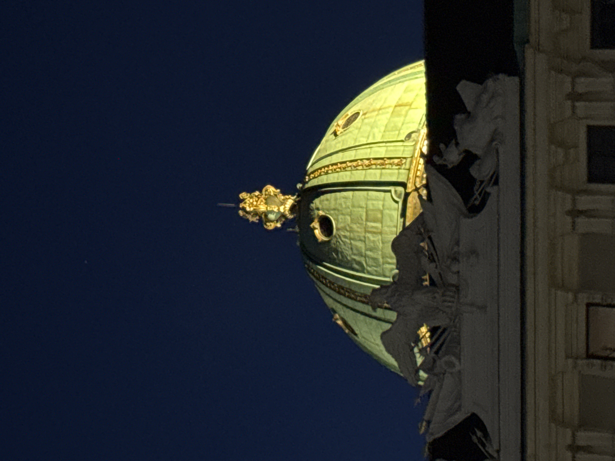 The image shows an upward-angled view of a green dome and a stone facade against a dark blue night sky. The dome is decorated with horizontal bands and circular windows. A golden, ornate architectural element extends from the side of the dome. The stone facade is textured and features sculptures that appear to be angels or mythical figures, adding to the grandeur of the architecture. The overall scene suggests a historical or religious building. The time of day is night, as indicated by the dark sky and the artificial lighting illuminating the dome. The lighting on the dome is a warm yellow, contrasting with the cool blue of the sky and the grey tones of the stone sculptures. The image is likely taken in Vienna, Austria, given the architectural style often found in European cities. There are no visible people or text in the image.