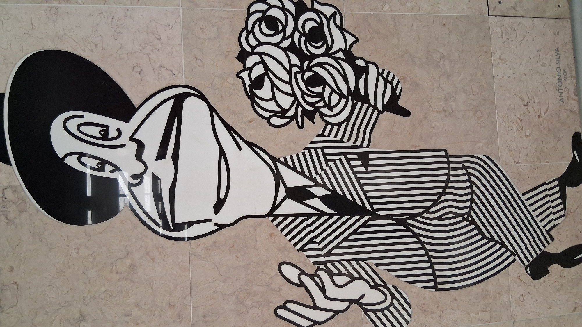 The image depicts a black and white mural on a tiled wall. The mural features a stylized cartoon character dressed in a pinstripe suit and hat, holding a bouquet of roses. The character has large eyes, a prominent nose, and an open mouth. One hand is outstretched, and the other is holding the flowers. The background is a light-colored, mottled tile. A signature "ANTONIO SILVA" with "ATOR" below it is visible in the lower right corner of the mural. This artwork is located in the Santa Justa Lift (Elevador de Santa Justa) in Lisbon, Portugal.