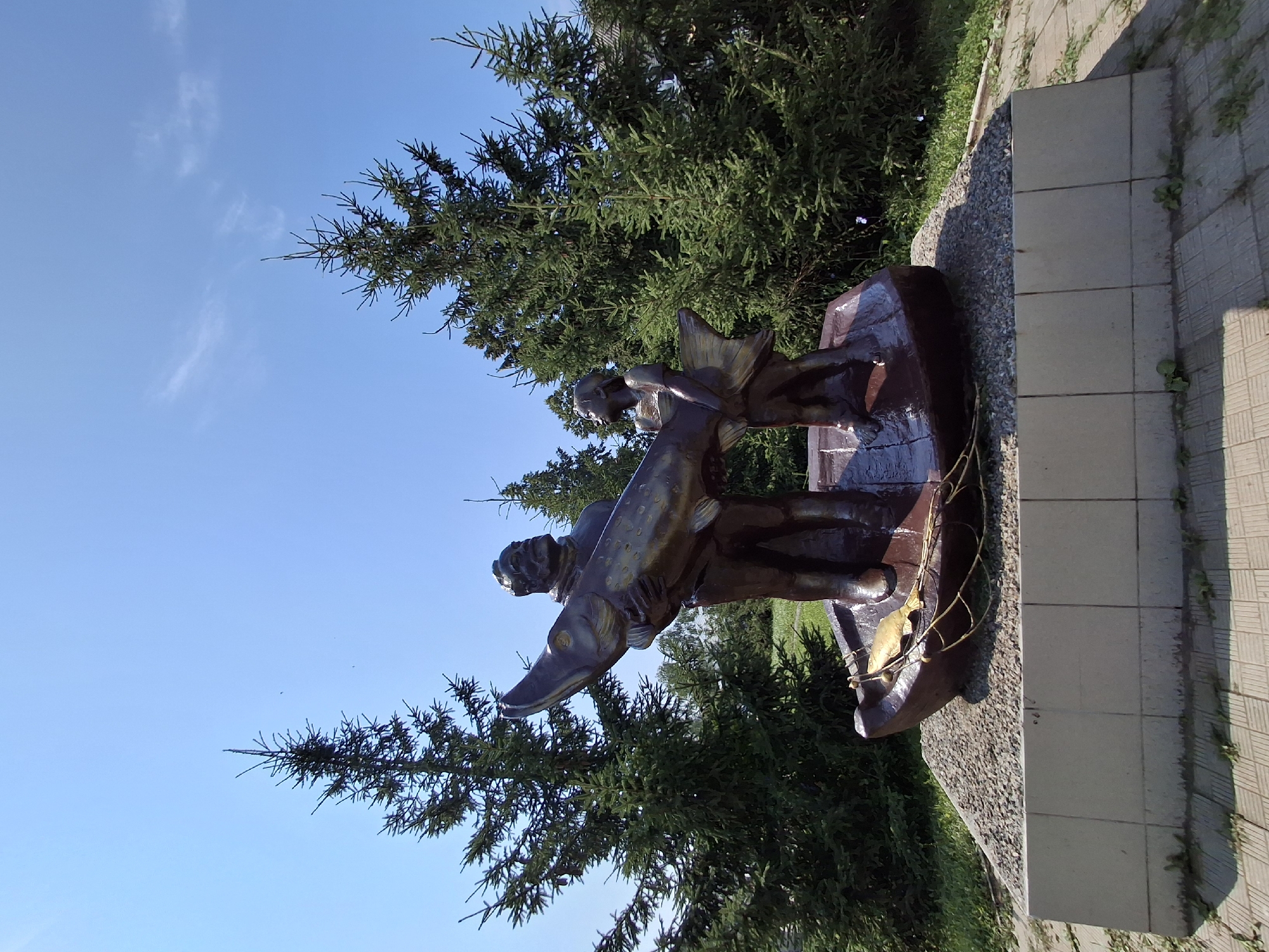 This image depicts a bronze sculpture of a man and a child in a boat, holding a large fish. The sculpture is situated on a bed of gravel, which is on top of a concrete pedestal. The pedestal is surrounded by a paved walkway. In the background, several tall fir trees are visible against a clear blue sky. The sculpture appears to be a monument or a landmark, potentially representing local fishing traditions. It is located in the city of Elovo, Russia.