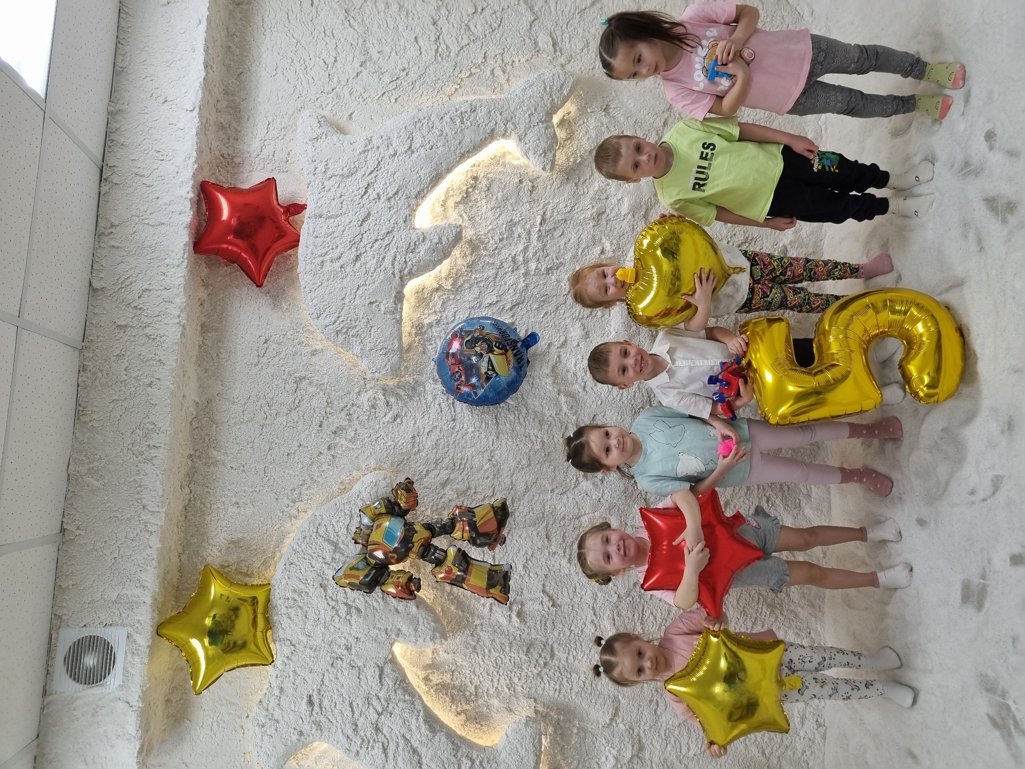 The scene is an indoor shot, possibly a playroom or party space, featuring seven children and several balloons against a textured, white wall.
The children are arranged in a row from top to bottom of the image. The top child is a girl wearing a pink shirt and gray pants. Next is a boy in a light green shirt with the word "RULES" on it, followed by a child holding a gold-colored balloon shaped like the number "2". Below that is a child in a white shirt holding a red object. The next child wears a light blue shirt and is holding a pink object. The child below this one is holding a red star balloon and wears a gray shirt and shorts. The last child holds a gold star balloon and wears a pink shirt and patterned pants.
Several balloons are present. A red star balloon is attached to the white wall in the upper left corner. A blue round balloon with cartoon figures is visible in the center of the wall. A cluster of Transformer-themed balloons is in the lower-left area of the wall. A gold star balloon is in the lower left corner, also attached to the wall. The children hold a gold "2", gold star, and a red star.
The environment features a textured, white wall with recessed lighting. A white fan is visible in the lower left corner of the image, indicating this is inside. The floor appears to be covered in a soft, white material.
The photograph may have been taken at the salt cave "SALT & KIDS" in the city of Chaikovsky, Russia.