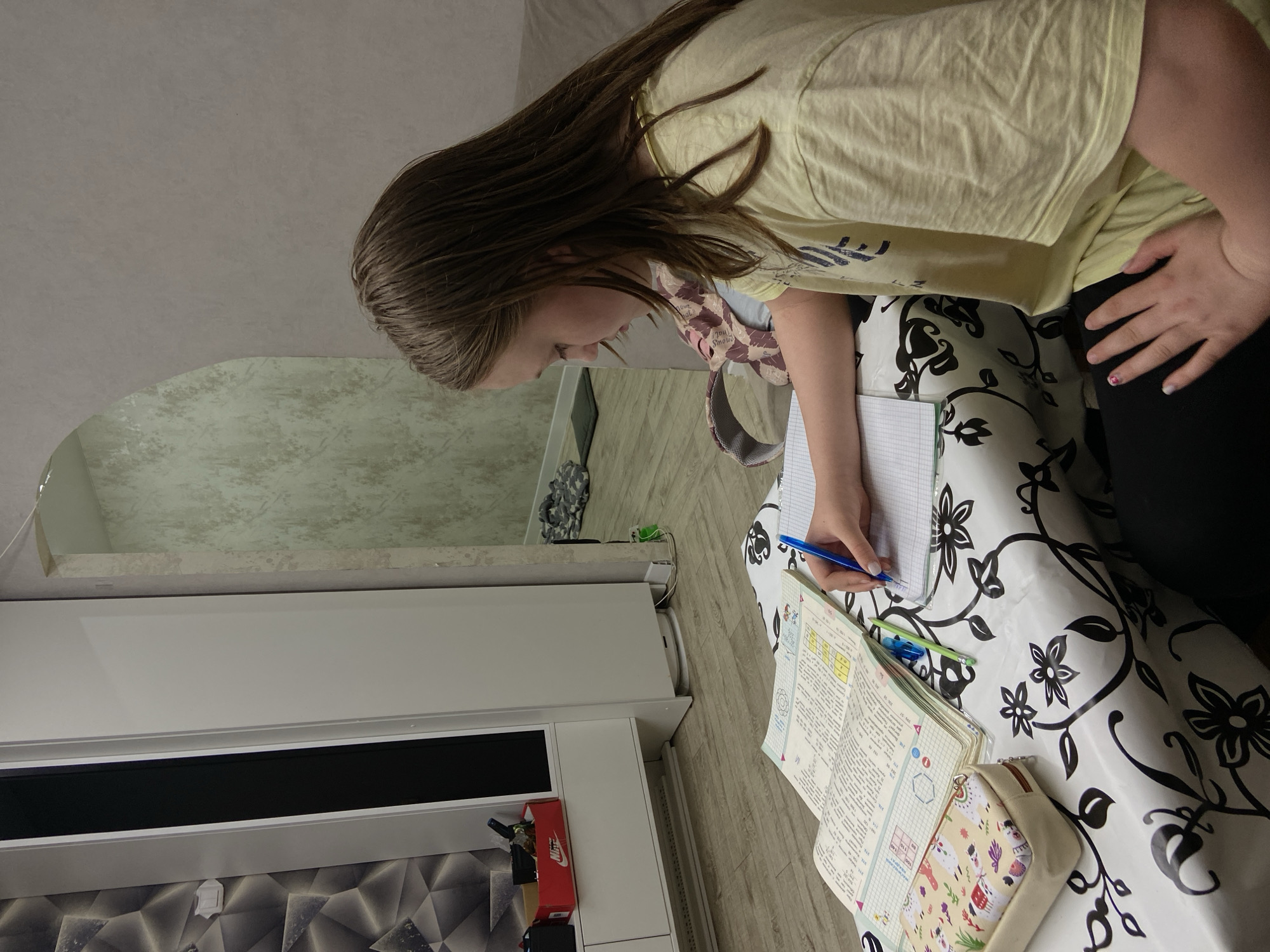 This image, taken in Chaikovsky, Russia, depicts a young person engrossed in studying indoors. The subject, likely a girl or young woman, has long brown hair and is wearing a light yellow short-sleeved t-shirt and dark pants. She is shown in profile, leaning over a table, actively writing in a notebook with a blue pen. Her expression suggests focus and concentration.
The table is draped with a white tablecloth featuring a distinct black floral and vine pattern. On the table, an open, grid-lined notebook contains handwritten notes, and an open textbook or workbook displays pages of Cyrillic text, numbers, diagrams, and what appears to be mathematical or scientific content, including a clear hexagonal shape. A cream-colored pencil case with playful llama or animal patterns is also present.
In the background, a modern white cabinet or media console with a black rectangular section is visible. Below it, the wall features a textured geometric pattern of triangles. Above the cabinet, a large mirror reflects a light, textured wall. The ceiling is a plain light grey, and the floor is light-colored wood or laminate. A red Nike shoe box can be seen on the cabinet, along with other small items and cables on the floor, and a camouflage-patterned object. One of the person's hands, resting on their hip, shows painted fingernails with different colors, including red and a lighter shade. The scene suggests a quiet moment of homework or personal study under ambient indoor lighting.