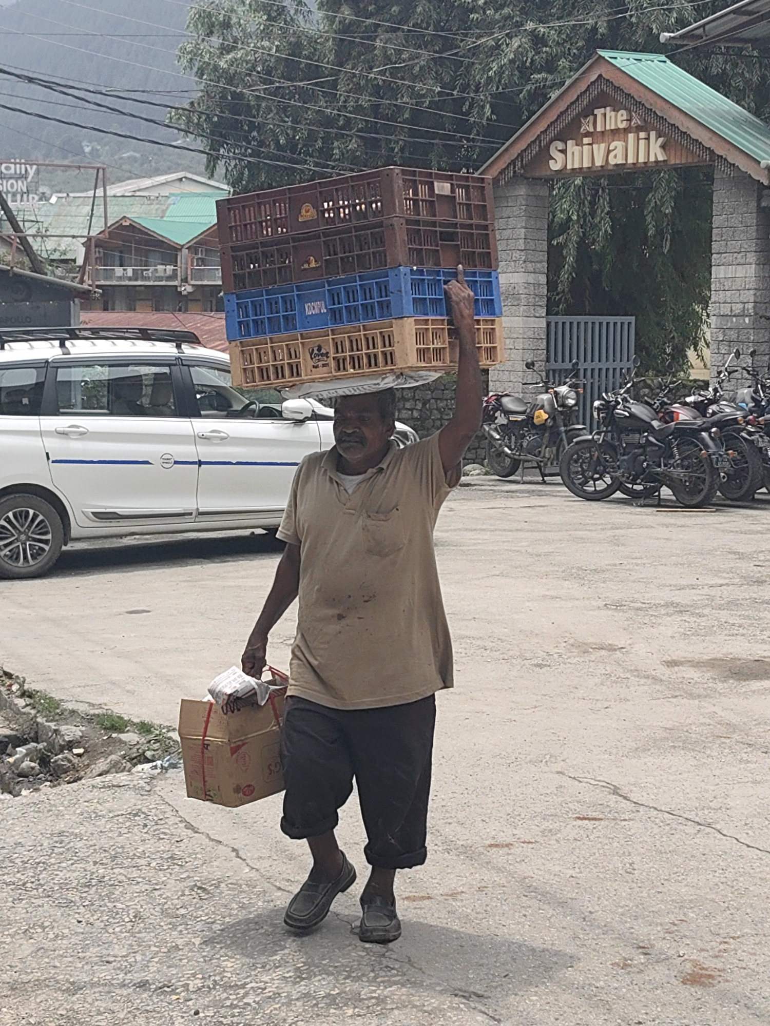 A man walks on a paved road in Manali, India, carrying a stack of plastic crates on his head and a box in his hand. He is wearing a tan polo shirt and dark pants. The crates are stacked three high, with the top one being brown, the middle one blue, and the bottom one tan. The box in his hand is made of cardboard and is tied with red string. In the background, there is a white car with a roof rack, several motorcycles parked, and a building with a sign that reads "The Shivalik". The area appears to be a commercial or tourist district, with some vegetation and mountains in the distance.