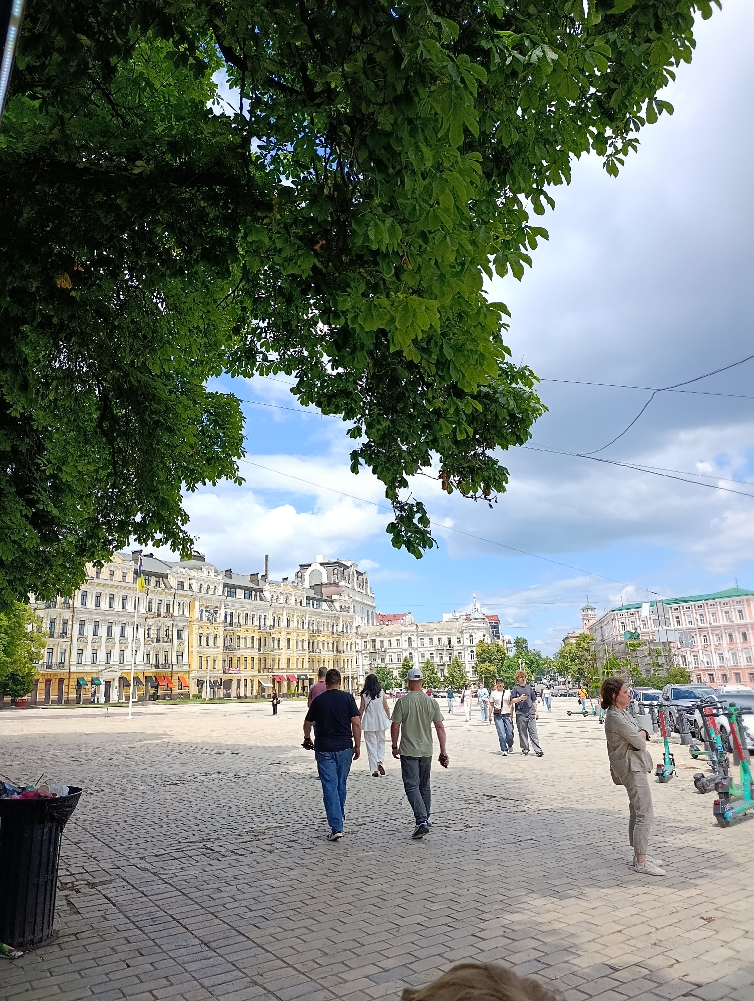 The image depicts a wide, paved square in Kyiv, Ukraine, likely in the Podil district, given the distinctive architecture. A large tree with lush green leaves dominates the upper left of the frame, its branches extending over the scene. In the background, a row of ornate, multi-story buildings with yellow and white facades line the square. Some of these buildings feature pitched roofs, visible balconies, and numerous windows. The sky above is partly cloudy with patches of blue.
In the foreground, a black trash can is visible on the left. Several people are walking across the paved area. A couple in the mid-ground walks away from the camera, with the man in a dark t-shirt and blue jeans, and the woman in a white outfit. To their right, another person in a green t-shirt and grey pants walks towards the background. Further to the right, a woman in a beige pantsuit stands with her arms crossed, leaning slightly. Several electric scooters are parked behind her. Other individuals and groups can be seen scattered throughout the square, walking or standing. The paving stones of the square are laid in a patterned brickwork. The overall atmosphere suggests a bright, possibly warm day in an urban setting.