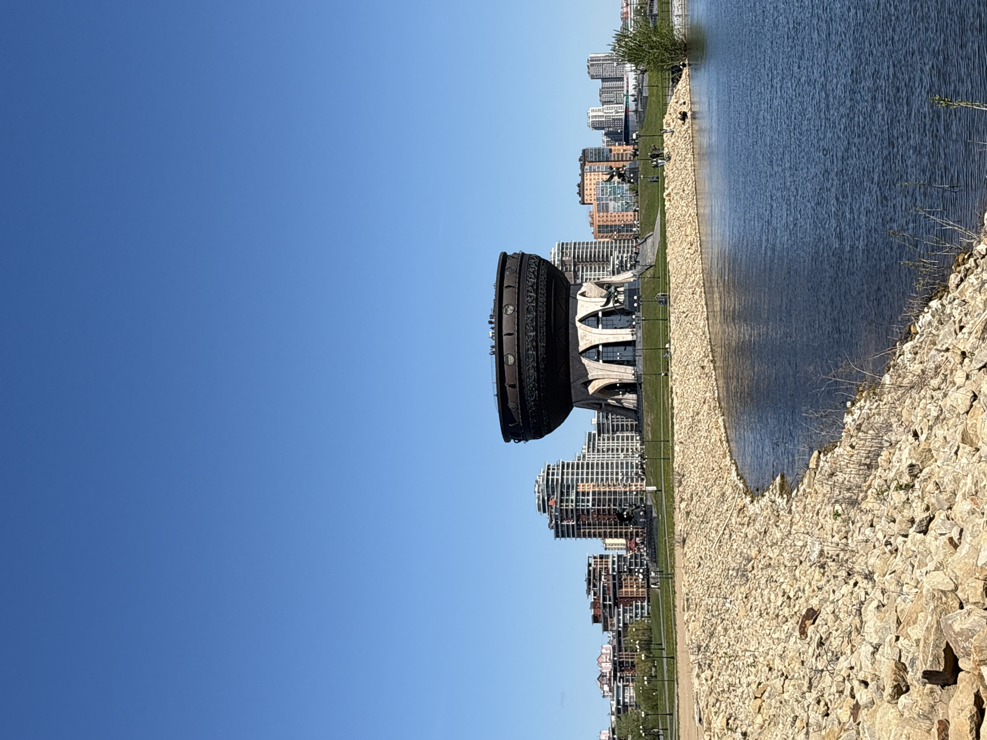 The image depicts the Marriage Palace Kazan, also known as the Kazan Family Center. It is a large cauldron-shaped building with an ornate exterior, situated on the bank of a river or lake, likely the Kazanka River. The structure is framed by modern high-rise buildings in the background. The shore is composed of rocks and gravel. The sky is a clear blue. The overall environment suggests a bright, sunny day.