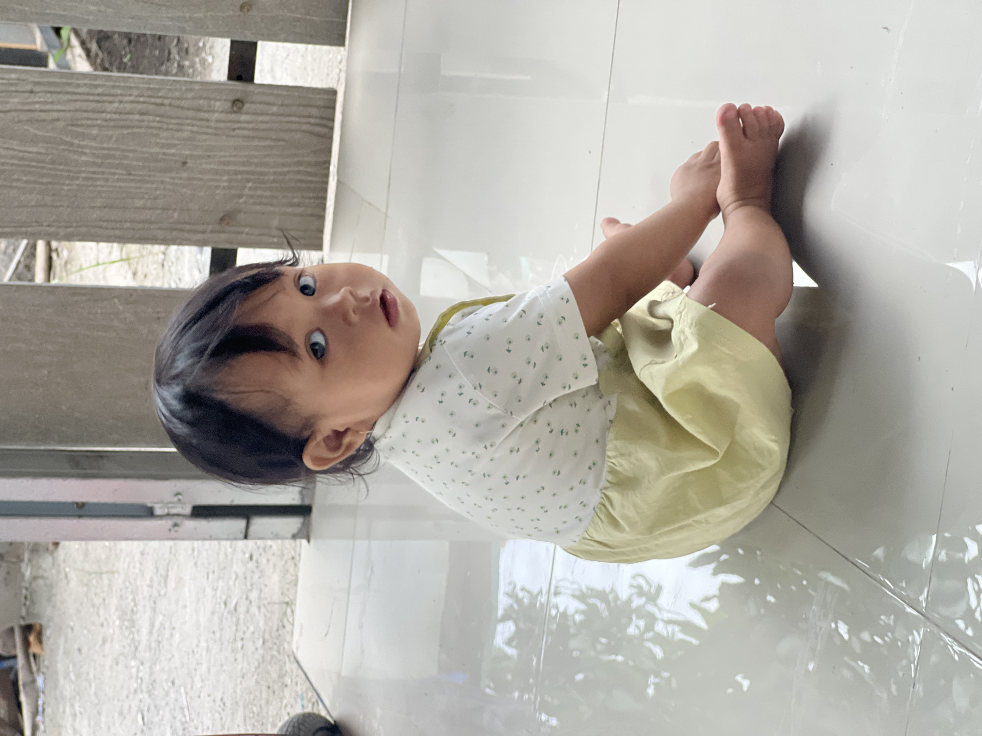 A young child with dark hair and light blue eyes is sitting on a shiny tiled floor. The child is wearing a white short-sleeved shirt with small green dots and a pale yellow ruffled skirt. The child is looking over their left shoulder towards the camera. The background shows a section of a grey wooden fence and a doorway leading outside to a gravel area. This scene appears to be an outdoor or semi-outdoor space within a residential area in Kecamatan Pati, Indonesia.