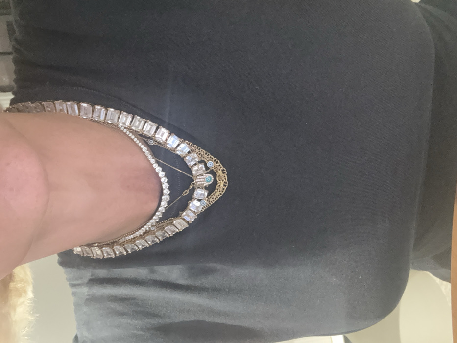 A close-up image depicts the upper torso and neck of an individual, likely female, wearing a black t-shirt. Light blonde hair is visible along the top-left edge of the frame. The subject's neck is adorned with multiple layered necklaces. The innermost necklace is a delicate chain featuring small, round, colorless stones. Directly beneath this, a more substantial necklace with large, emerald-cut clear stones set in a rectangular pattern is visible. Interspersed are several thinner, gold-toned chains: one includes small blue evil eye beads, another features a central Hamsa hand pendant with a blue stone, and a third displays an intricate, possibly filigree, chain design. The background is out of focus, showing blurred light-colored surfaces consistent with an indoor setting. The specific location within Dubai, United Arab Emirates, cannot be determined from the visual content provided.