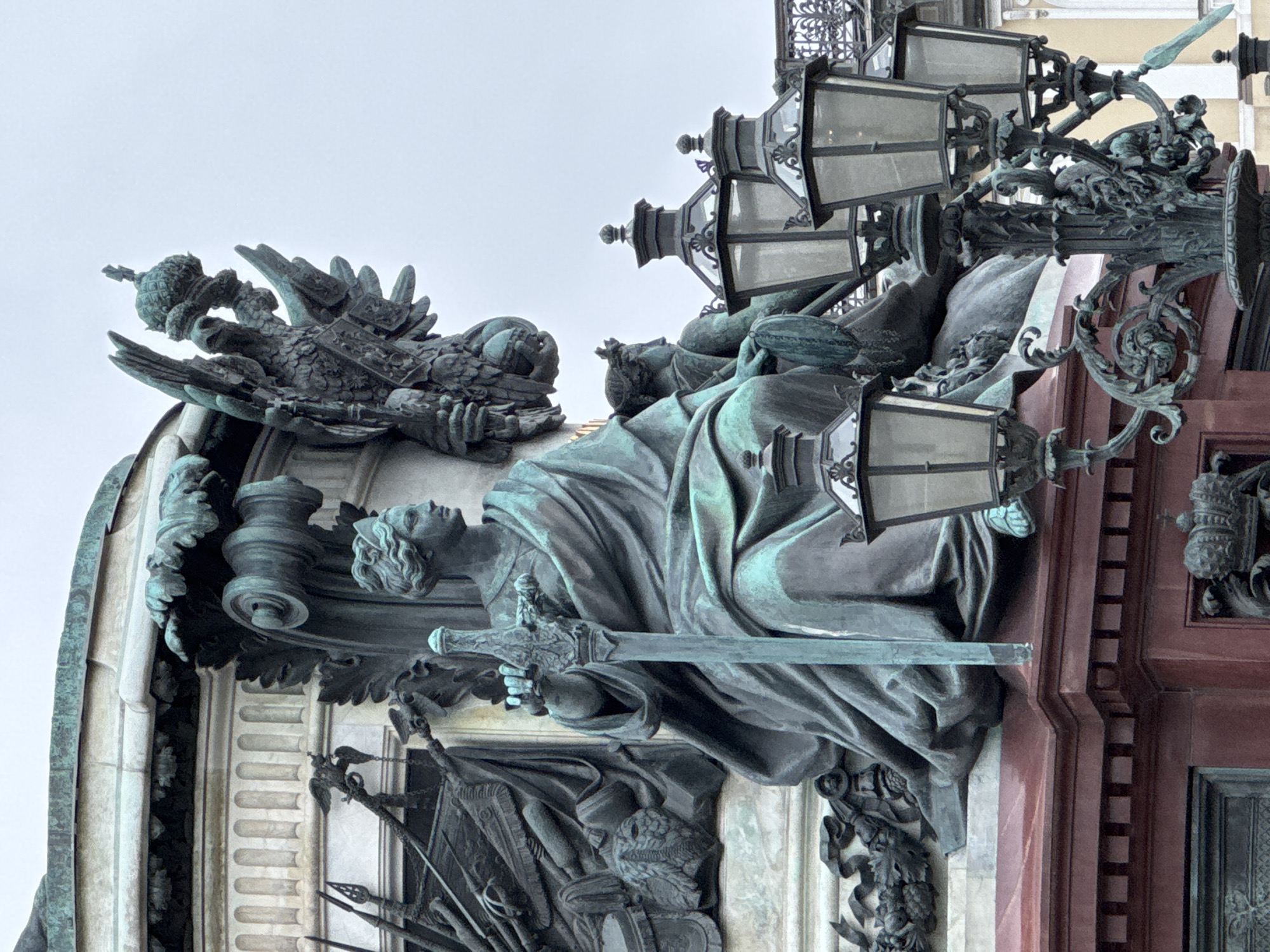 This image captures a detailed, weathered bronze sculpture, characterized by a prominent blue-green verdigris patina. The central element is a draped figure, possibly allegorical, holding a large, ornate sword vertically with both hands. The figure's face is serene, looking upwards.
Above and to the left of this figure, a majestic two-headed eagle is depicted, clutching an imperial orb and scepter. This symbol strongly indicates Russian imperial heritage. The sculpture is flanked by an array of elaborate lanterns featuring multiple glass panes and intricate scrolled metalwork, suggesting they are functional lighting elements on a grand scale.
The sculpture is integrated into a larger architectural structure, evidenced by fluted columns and a substantial base or plinth, which appears to be made of a dark reddish-brown material. Below the main figure and the sword, relief sculptures portray military armaments such as helmets, shields, and spears, suggesting themes of defense, victory, or historical significance.
The scene is set outdoors during the daytime, with a clear, light sky providing soft, diffuse lighting that highlights the intricate details and texture of the bronze. The overall impression is that of a significant historical or commemorative monument, characteristic of the grand public art found in Saint Petersburg, Russia. No visible text is discernable in the image.