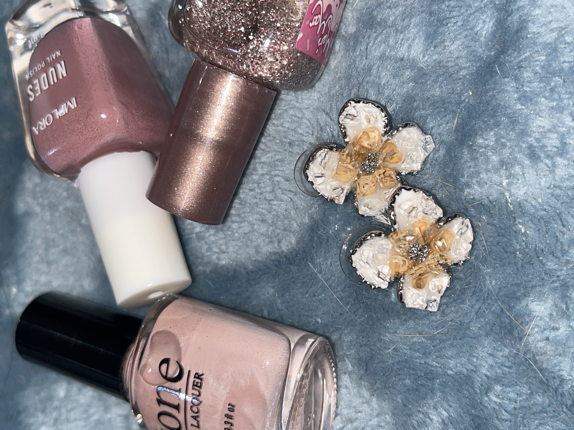 The image captures a still life arrangement of beauty products and accessories on a soft, blue, fuzzy fabric surface, possibly a blanket or towel.
The main subjects are three bottles of nail polish and two decorative flower-shaped objects.
One nail polish bottle, prominent in the upper left, is a dusty rose or nude color. Its label clearly reads "IMPLORA NUDES NAIL POLISH." It has a white cap.
Adjacent to it, towards the top right, is a glittery nail polish bottle in a rose gold or copper hue. Its cap has a metallic, shimmering finish. A partial label on this bottle shows fragmented text that is difficult to decipher fully.
In the bottom left corner, another nail polish bottle is visible. This one is a pale pink or nude shade with a black cap. The visible text on its label includes "one LACQUER" and a volume measurement, likely "3 fl oz" though blurry.
To the right of the nail polish bottles, two identical flower-shaped objects are displayed. These objects appear to be decorative, possibly earrings or embellishments. Each flower has several white, almost translucent petals, with a central cluster of small, golden or amber-colored stones or beads. The very center of each flower features tiny, sparkling silver elements, resembling glitter. The overall design suggests a delicate and somewhat intricate craftsmanship.
The background fabric is a solid, medium blue color with a plush, textured surface. Several thin strands of hair are visible on this surface, suggesting a casual, lived-in environment. The lighting is even, illuminating the items without harsh shadows, likely from an indoor source. No people are visible, and there are no specific cues for time of day or weather. The arrangement appears to be a casual display of personal care and adornment items.