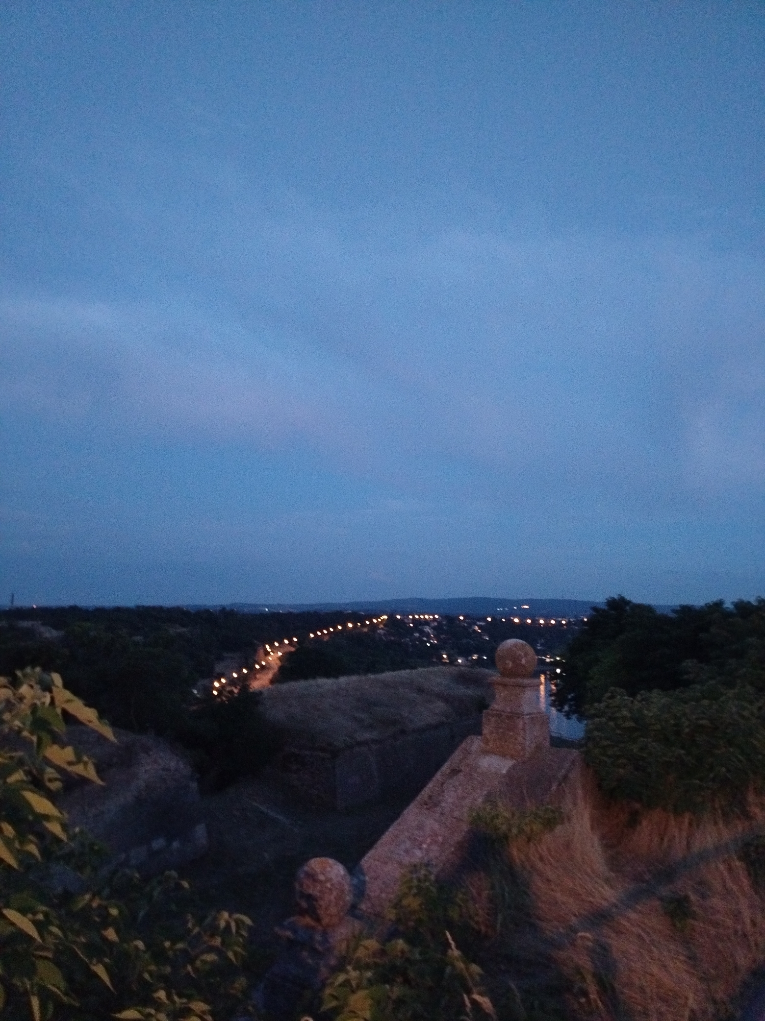 The image captures a panoramic view of Petrovaradin, Serbia, at dusk. The sky is a deep blue, transitioning from lighter hues at the horizon to a darker shade overhead. In the distance, a road lined with streetlights winds through the city, casting a warm glow. The landscape below consists of dark green foliage and structures, suggesting a hilly or elevated terrain. In the foreground, a stone structure, possibly part of a fortification, is visible, covered with dry grass. A small, round finial adorns the top of the stone structure. The overall scene conveys a sense of calm and twilight in the historic city.