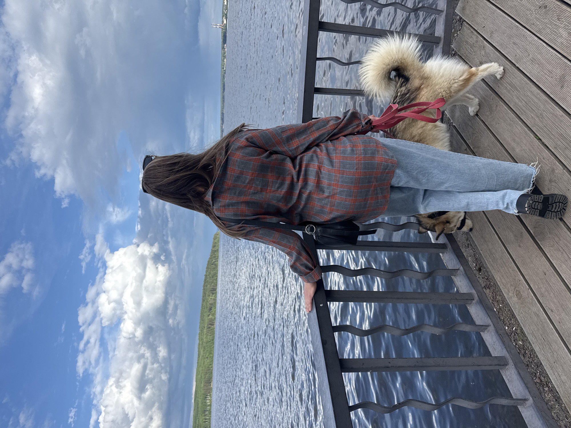 A woman and a dog stand on a wooden platform looking out over the water, possibly on the Kama River. The woman has long brown hair and wears sunglasses, a gray and orange plaid shirt, and blue jeans. She leans against a dark metal railing with her right hand. A black bag hangs from her shoulder. The dog, which is medium in size with long brown and white fur, stands next to the woman with a red leash attached to its collar. The river spans the horizon and a city skyline is visible in the distance. The sky is blue with patches of white clouds. The image suggests a daytime scene near the city of Chaikovsky, Russia, given the potential identification of the Kama River.