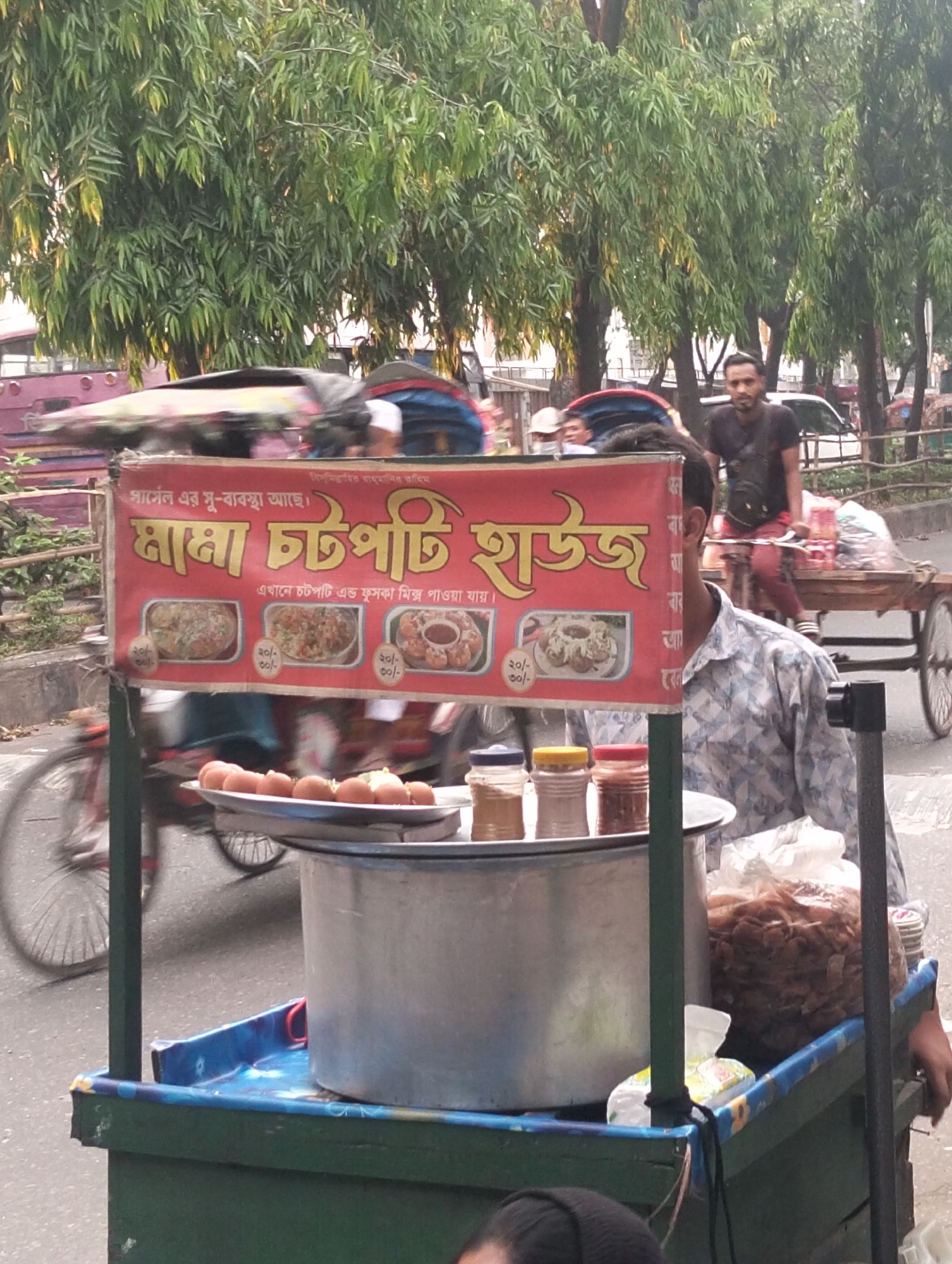 The image shows a street food vendor's cart in Dhaka, Bangladesh. The cart is selling "chatpati," a popular South Asian street food. A sign on the cart reads "Mama Chatpati House" in Bengali and advertises that chatpati and phuchka mix are available for 20-30 taka. The cart also has eggs, spices, and what appears to be fried chips in bags. A vendor is standing near the cart. In the background, there is street traffic, including a rickshaw puller and other pedestrians, trees, and buildings, suggesting a busy urban environment.