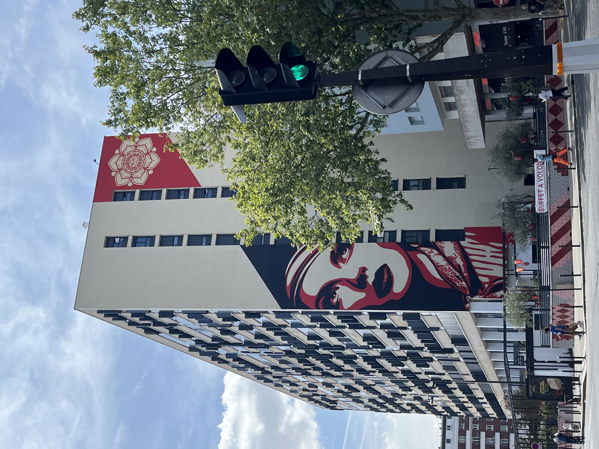 The image depicts a large mural on the side of a modern apartment building in Paris, France. The mural features a stylized portrait of a woman with striking red lips and eyes, rendered in a bold, graphic style, alongside a red geometric pattern with a lotus-like symbol at the top of the building. The building itself is tall and rectangular, with numerous windows. In the foreground, a traffic light shows a green signal, and trees with lush green foliage partially obscure the building. The street level shows some construction barriers, storefronts, and a few pedestrians walking along the sidewalk. One pedestrian is wearing bright orange pants and a blue shirt, and another is wearing a white shirt and dark pants. The sky above is partly cloudy.