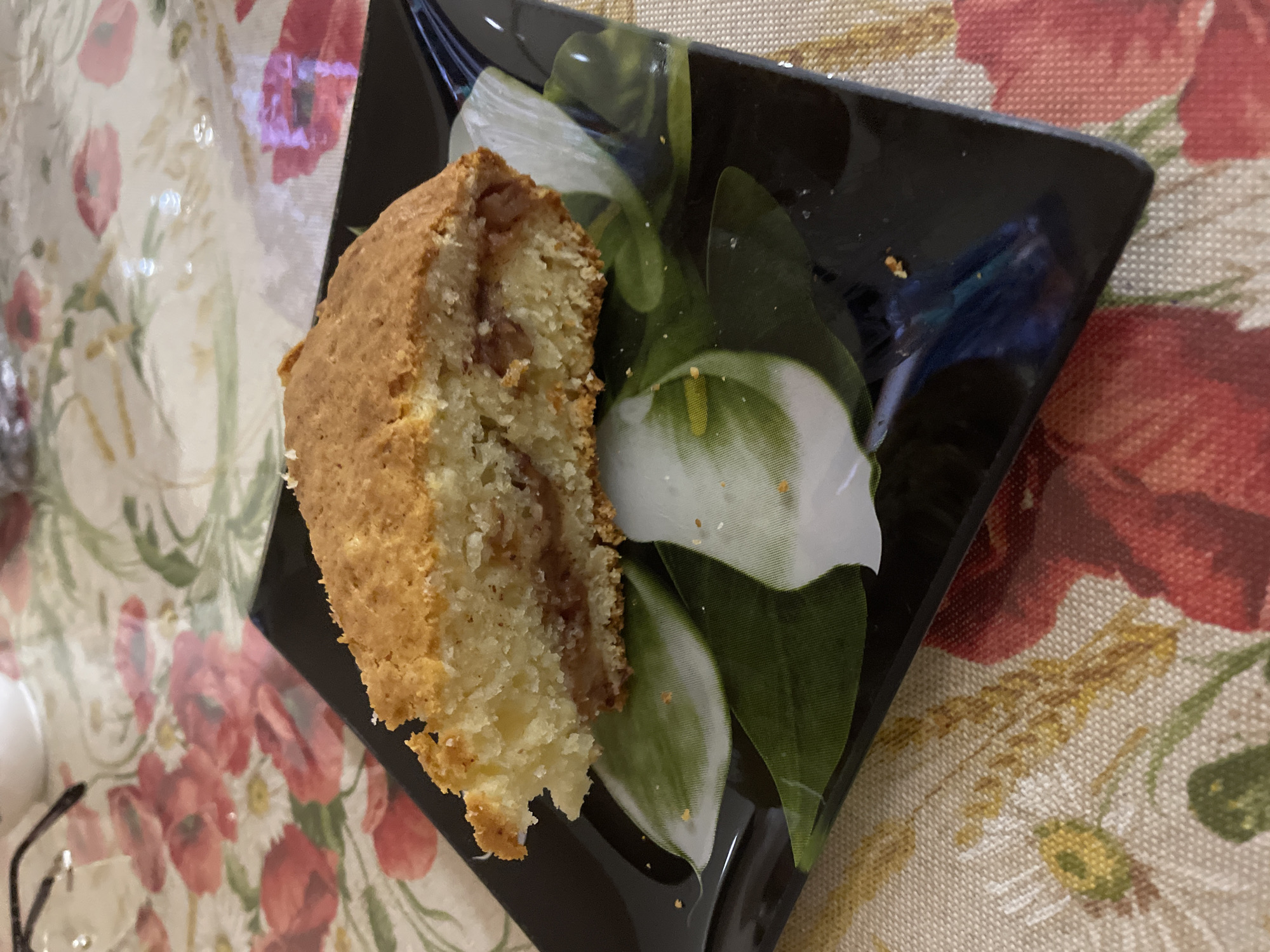 A slice of cake sits on a square black plate decorated with white lilies and green leaves. The cake has a light brown crust and a filling, appearing to be on a tablecloth with a floral pattern, featuring red poppies and wheat on a beige background. Eyeglasses are partially visible in the lower-left corner of the image. The location is specified as Moskovskiy, Russia.