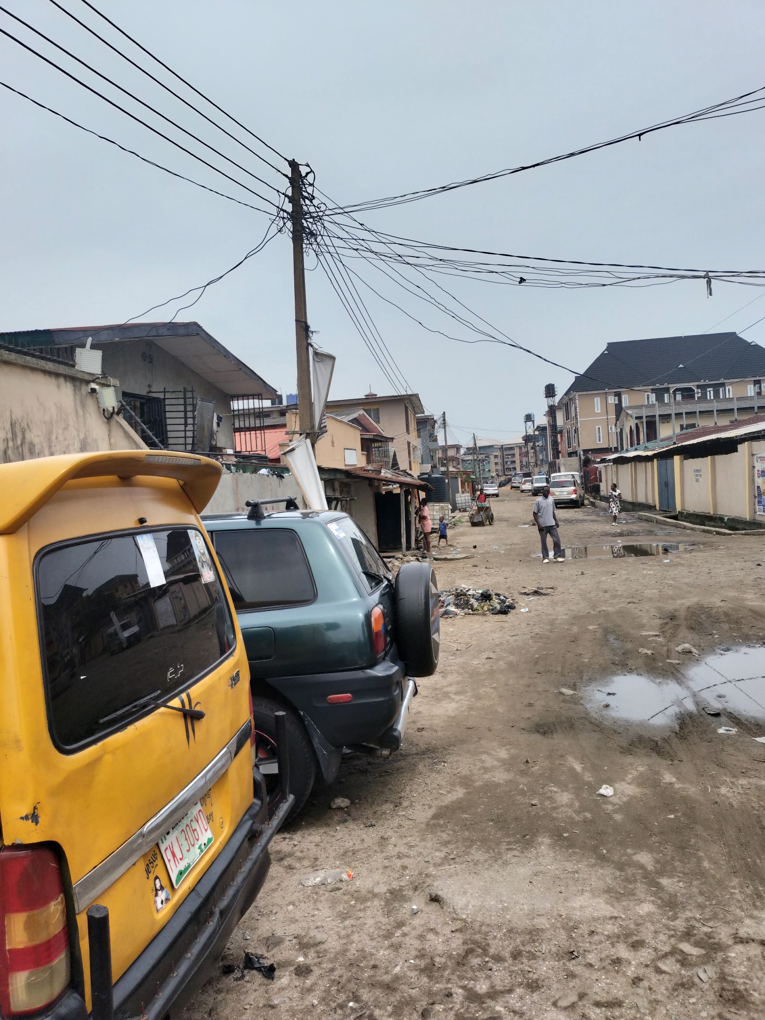 The image depicts a street scene in Alaiyabiagba, Nigeria. The unpaved road is muddy and potholed, with standing water. A yellow minibus taxi and a green SUV are parked on the left side of the road. Overhead power lines are visible, attached to a weathered pole. In the background, people are walking along the road. Buildings of varying sizes and conditions line the street. There is debris and litter scattered in the street.