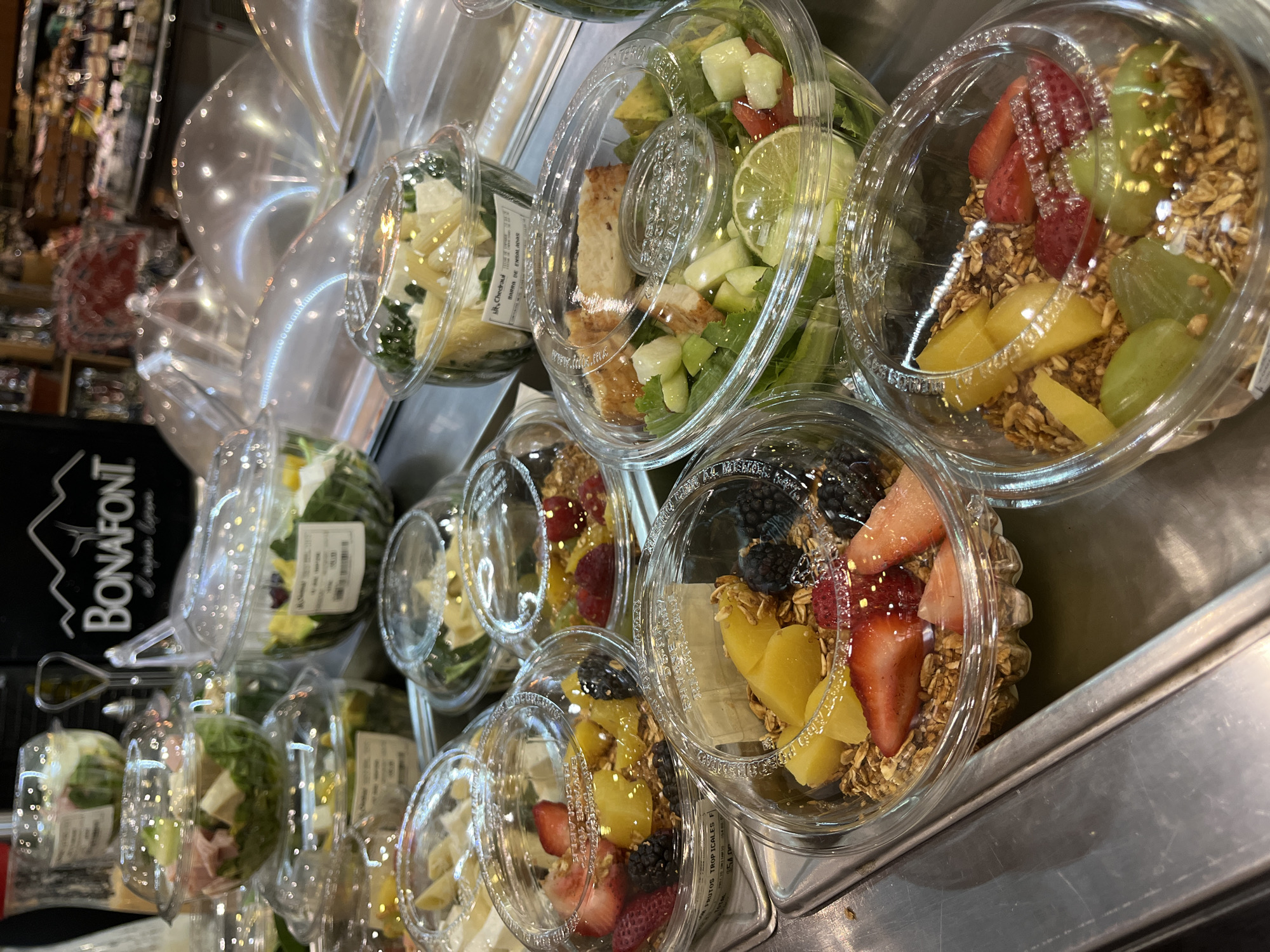 The image depicts a display of pre-packaged salads and fruit bowls in clear plastic containers, presumably for sale. The containers are arranged on a stainless steel shelf or surface. The food items include various salads with leafy greens, cheese, and what appears to be grilled chicken, as well as fruit bowls containing granola, mango, strawberries, blackberries, and grapes. A sign with the brand name "BONAFONT" is visible in the background. The specific location in San Bernardino Tlaxcalancingo, Mexico cannot be determined from the image.