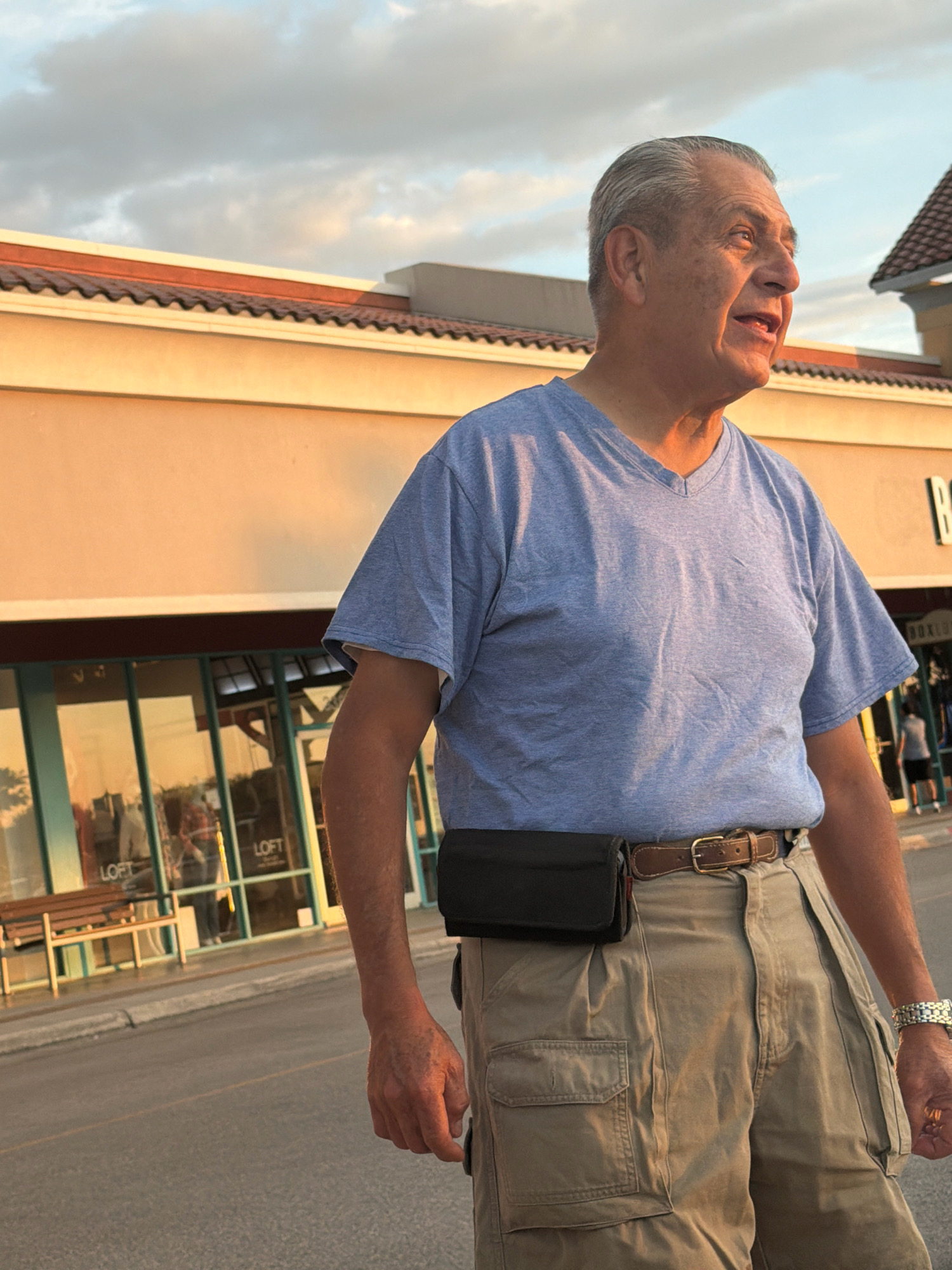 An older man with gray hair and a light blue t-shirt stands on a paved surface in front of a building with a tan facade and a brown tiled roof. He is wearing tan cargo pants and a brown belt with a black fanny pack attached. The building has large windows, and the word "LOFT" is visible through them. A wooden bench is situated outside the building. The sky above is cloudy with a golden hue, suggesting it is either sunrise or sunset. The scene appears to be in a commercial area.