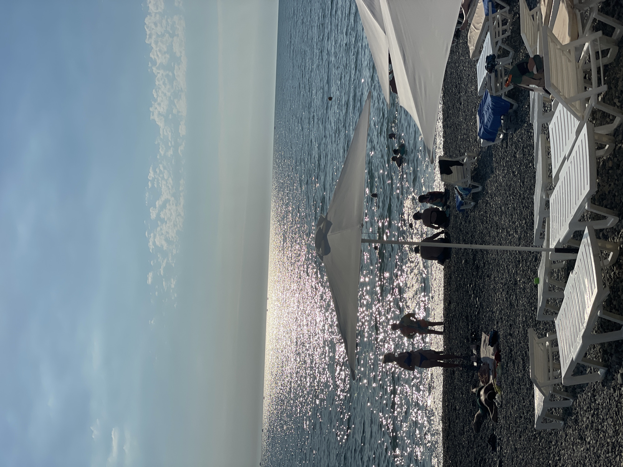 The image depicts a sun-drenched pebble beach in Sochi, Russia, with the Black Sea stretching out to the horizon. The water is illuminated by the sun's reflection, creating a shimmering effect. Several people are visible, some wading in the shallows and others relaxing on the beach. White beach umbrellas and lounge chairs are scattered across the pebbles, suggesting a popular tourist spot. In the foreground, lounge chairs are neatly arranged, some with blue towels laid out on them. A woman in a blue bikini and a man in colorful swim trunks stand near the water's edge, silhouetted against the bright sea. Further back, a person lies on a towel on the beach, and other beachgoers are in the water. The sky is a light blue with scattered clouds. The scene captures a typical summer day at a Black Sea resort.