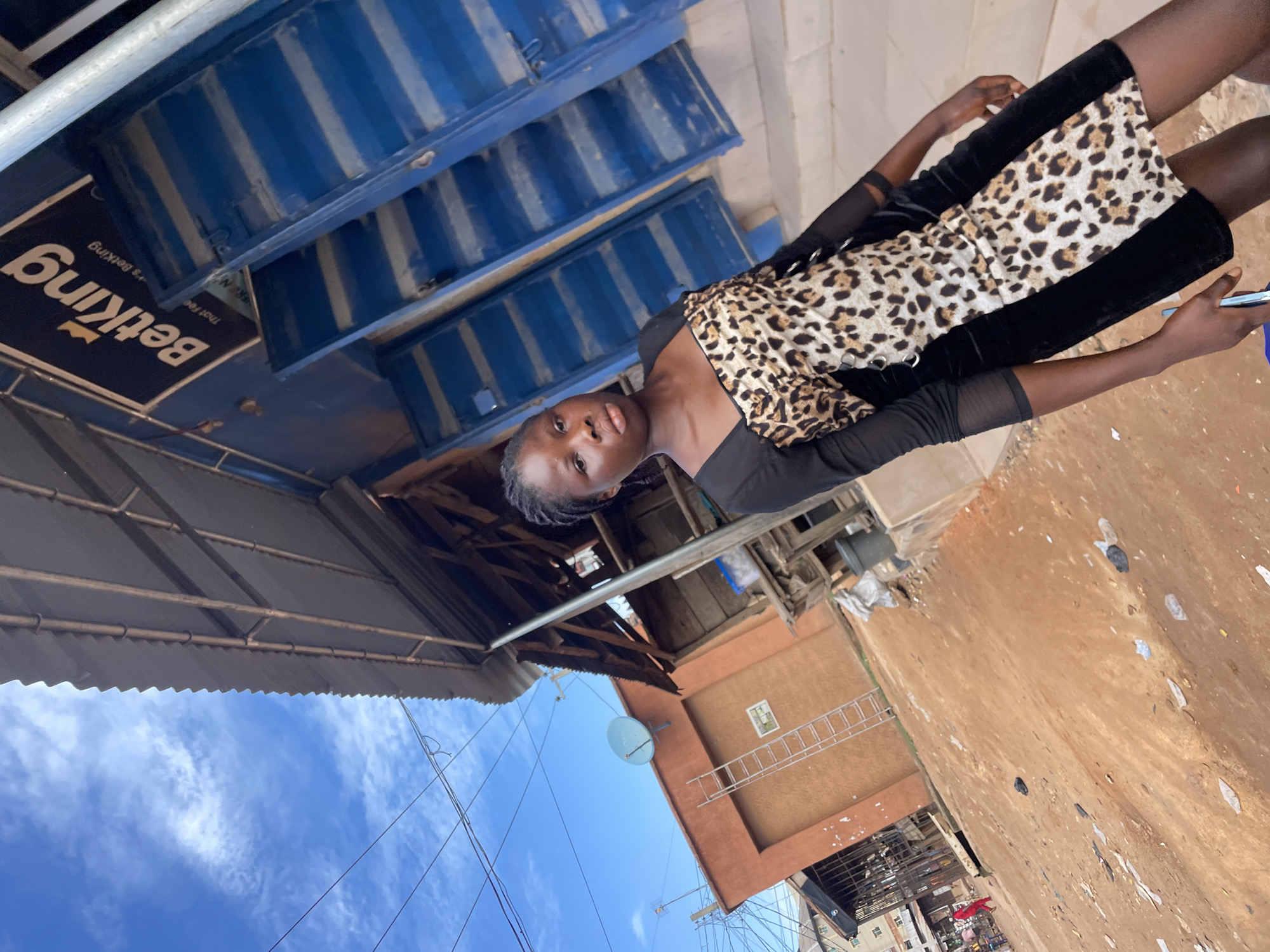 A young woman is the focal point of this ground-level photo, appearing to be outdoors in Onitsha, Nigeria. She is wearing a black top with sheer sleeves and a leopard-print skirt with black velvet panels. She holds a mobile phone in her left hand.
Overhead, there is a metal awning that partially covers the area. There is a blue structure with stacked storage boxes behind the woman and a sign that says "Betking." The ground is dirt and there's debris and scattered papers present. A building is visible in the background with a satellite dish attached to it. The sky above the building is clear and blue. A ladder rests against the building's side. There are utility wires stretching across the sky.