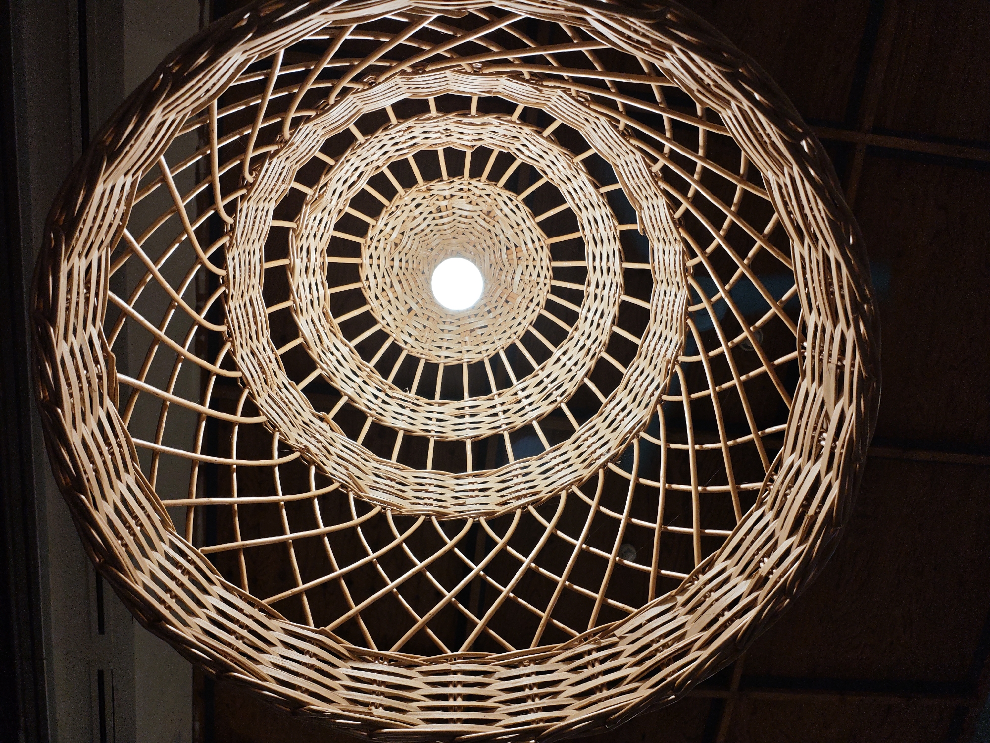 The image shows a close-up, low-angle shot of a woven rattan lampshade, illuminated from within by a bright light bulb. The lampshade is spherical with a concentric, spiral pattern. The woven material creates a textured surface with numerous openings that allow light to pass through. The background is dark and out of focus, with hints of what appears to be a ceiling with visible rafters or structural elements. The scene is indoors, likely in a room or dwelling. There is no information in the image to suggest it is located in Alushta, Ukraine.