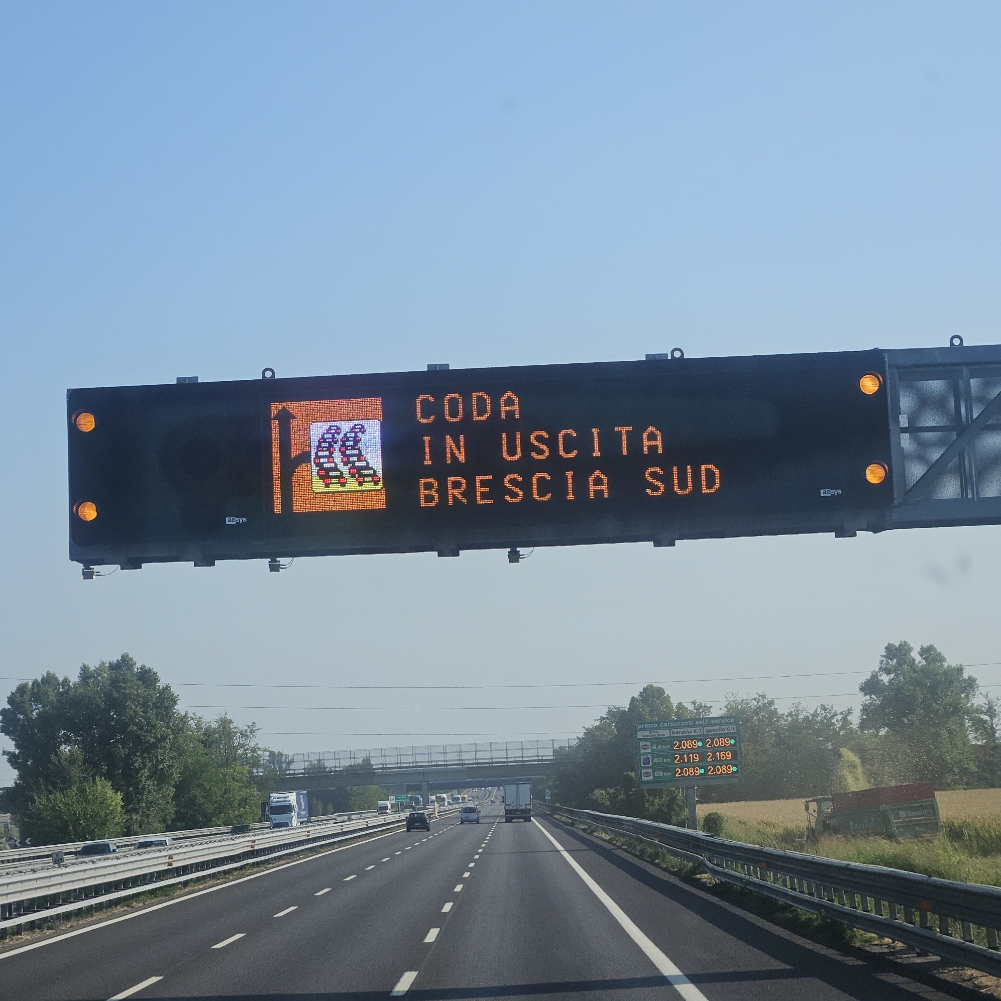 This media file captures a highway scene in San Zeno Naviglio, Italy, on a clear, sunny day. The primary focus is an overhead electronic traffic sign displaying a message in orange LED lights. The sign reads "CODA IN USCITA BRESCIA SUD," which translates to "Traffic Jam at Exit Brescia South." To the left of the text, a graphic depicts a queue of cars, further reinforcing the message of congestion.
Below the traffic sign, another electronic display provides fuel prices at various distances. It lists "prezzo carburanti self-service" (self-service fuel prices) and shows prices for "benzina" (gasoline) and "gasolio" (diesel) at 4.6 km, 40 km, and 69 km distances. For instance, at 4.6 km, both gasoline and diesel are priced at €2.089 per liter.
The highway itself is visible, with multiple lanes and standard white lane markings. Cars and trucks are driving on the road, though the traffic sign suggests potential delays ahead. The surrounding environment features lush green trees and vegetation, with a field of crops and agricultural machinery, including a tractor with a trailer, visible to the right of the highway. The bright sunlight and clear blue sky indicate fair weather. The overall impression is one of normal highway travel interrupted by a traffic issue, with practical information about fuel prices also being provided to drivers.