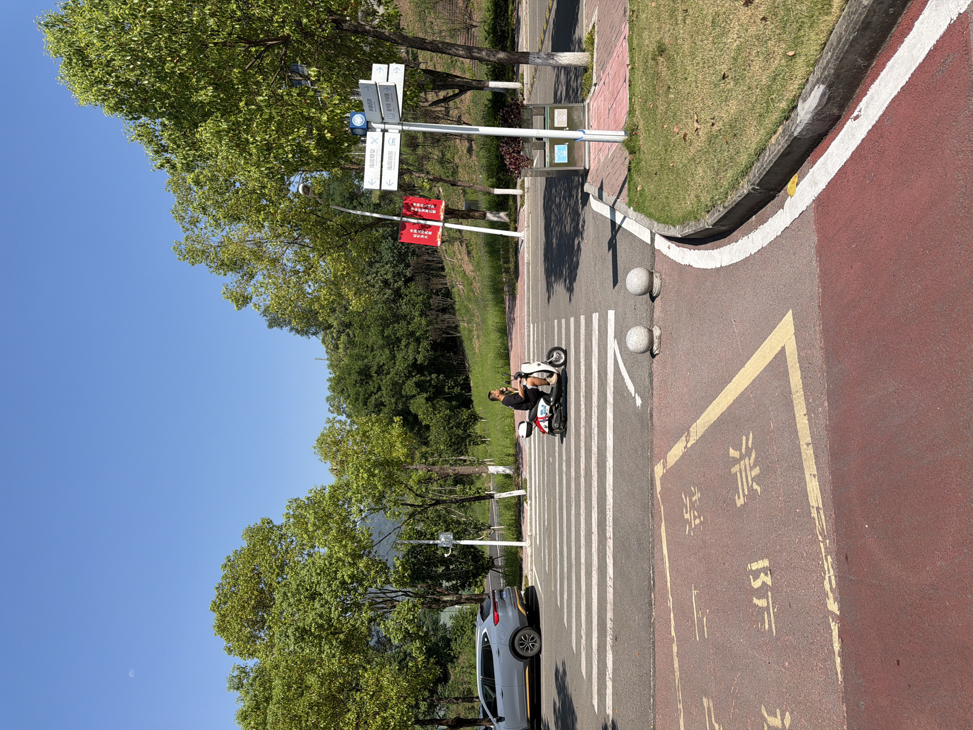 A young Asian man rides a white electric scooter across a pedestrian crossing in Jiangjin, China. He is wearing a black t-shirt and shorts. The scooter has a white and red body and a small white box on the back. The pedestrian crossing is a series of white stripes on asphalt. To the left of the crossing, a gray SUV is parked. Behind the crossing, there are trees and a grassy hillside. A street sign with Chinese characters and arrows is visible on the right side of the image, pointing towards different locations. A trash can is located near the street sign. The sky is clear and blue, and the sun is shining. The overall scene depicts a typical street view in a suburban or semi-urban area of China.