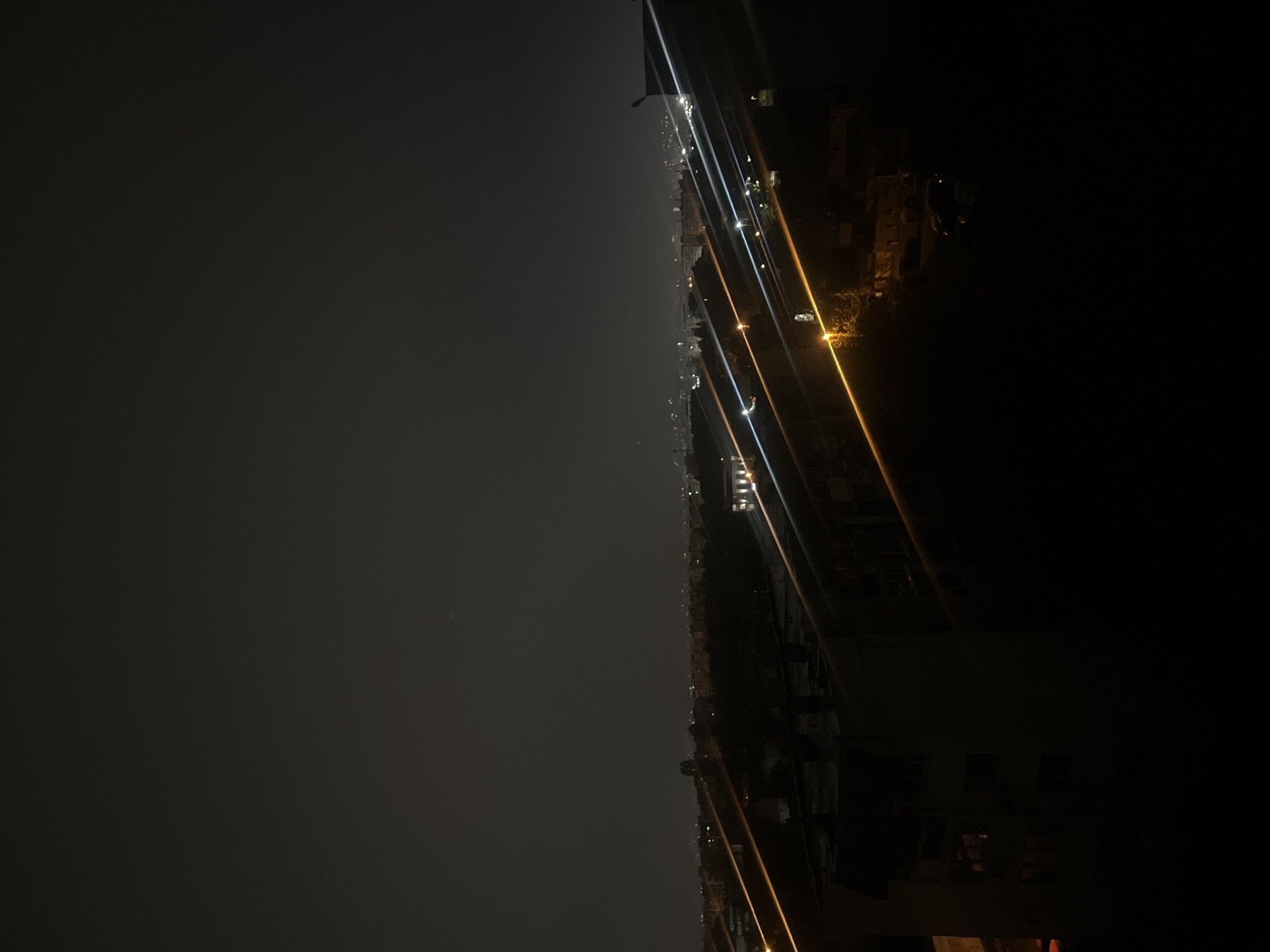 This media file presents a panoramic nighttime view of a city, likely Sofia, Bulgaria, captured from a high vantage point. The image depicts a vast urban landscape illuminated by artificial lights under a dark, clear sky.
The sky occupies the upper portion of the frame, appearing as a deep, gradient gray to black, devoid of clouds or significant celestial objects, suggesting a clear night. A faint, almost imperceptible light source or distant star might be visible in the upper left.
The mid-ground reveals the sprawling city, characterized by numerous scattered lights that mark the presence of buildings and infrastructure. These lights create a horizontal band across the image, appearing as bright, sometimes streaky, points and lines of light. The dominant colors of these urban lights are cool whites and warm yellows or oranges, indicating various types of streetlights, building illumination, and possibly vehicle head/tail lights from a distance. Individual buildings are mostly indistinct due to the darkness and distance, dissolving into a shimmering tapestry of light.
In the immediate foreground, particularly on the right side, some closer structures or parts of buildings are visible as dark silhouettes. These areas also feature pronounced light streaks and flares, predominantly in bright white and golden-yellow hues, running diagonally across the lower and upper right sections. These suggest bright artificial light sources close to the camera, possibly streetlights or overhead cables, causing a lens flare effect.
No people or specific activities are discernible in the image, which focuses on the general ambiance of a city after dark. The scene conveys a sense of quiet vastness and the enduring glow of urban life. There is no visible text within the image. The time of day is clearly night, with no apparent adverse weather conditions such as fog or rain, allowing for a relatively clear view of the distant lights.