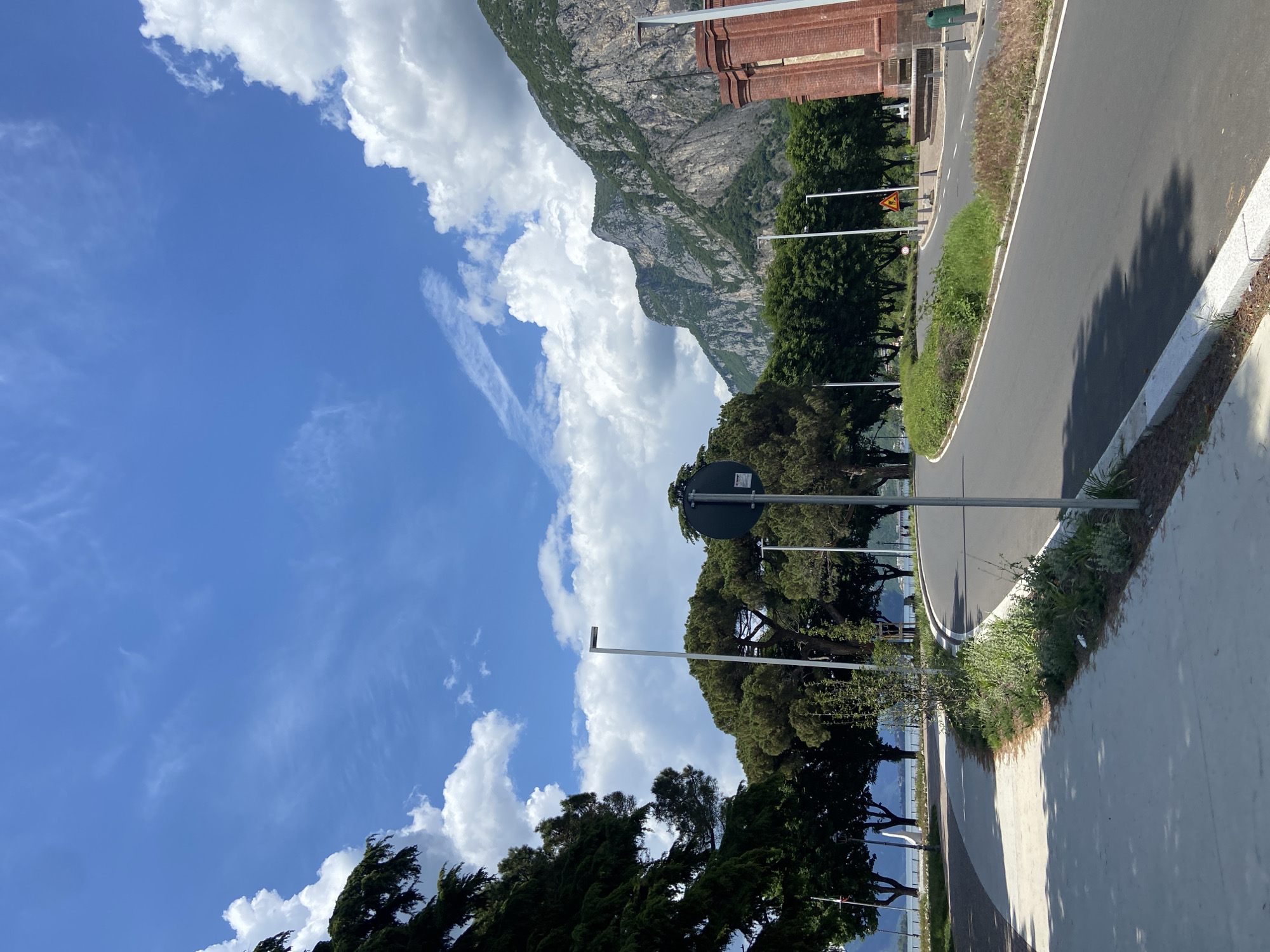 The image shows a view of the city of Lecco, Italy. The perspective is from a slightly elevated position, looking towards a mountain range in the background. In the foreground, a road curves along the coastline. The sky is blue with scattered cumulus clouds. Along the road are trees, lampposts, and various plants. A sidewalk runs parallel to the road. A building with brick architecture is visible on the right side of the image, adjacent to the mountain. No people are visible.
