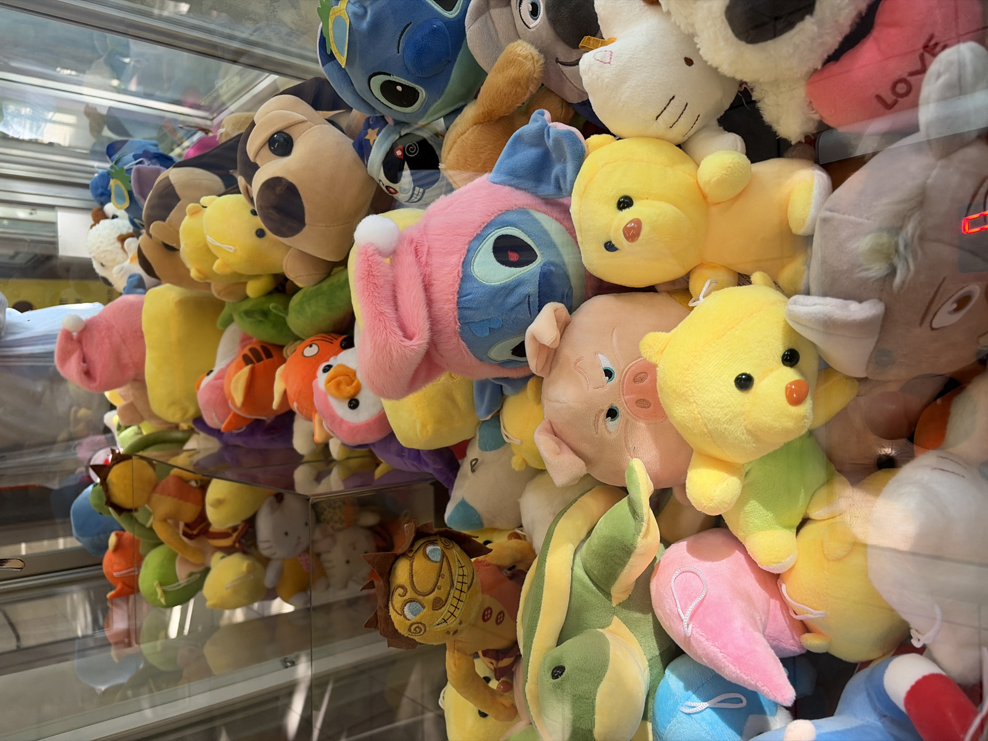 This vibrant image captures a dense collection of plush toys tightly packed inside a glass claw machine, likely found in an amusement arcade or similar entertainment venue in Sochi, Russia. Hundreds of soft, colorful characters are crammed together, forming a jumbled landscape of potential prizes.
Among the prominent subjects are multiple yellow teddy bears or chicks, some with rosy cheeks, and a large blue plush of Stitch from Lilo & Stitch, partially obscured by a pink version of the same character or a similarly shaped creature. Other visible toys include a pink pig, a brown and black spotted dog-like plush, a white Hello Kitty-style toy, an orange tiger, a green and yellow snake or dragon, and a distinctive brown lion or monster character with a wide, grinning mouth and large eyes. A smaller, heart-shaped pink plush with the word "LOVE" in white lettering is also visible in the upper right section of the display.
The setting is brightly lit, with artificial lights reflecting off the clear glass panels of the machine, creating subtle distortions and showing faint reflections of the surrounding environment. The chaotic arrangement of the toys suggests they are awaiting a player to skillfully maneuver the crane to win a prize. The image is taken from a slightly elevated perspective, looking down into the machine, highlighting the sheer volume and variety of the stuffed animals available. The abundance of vibrant colors – including shades of yellow, pink, blue, green, and brown – contributes to a lively and inviting atmosphere.