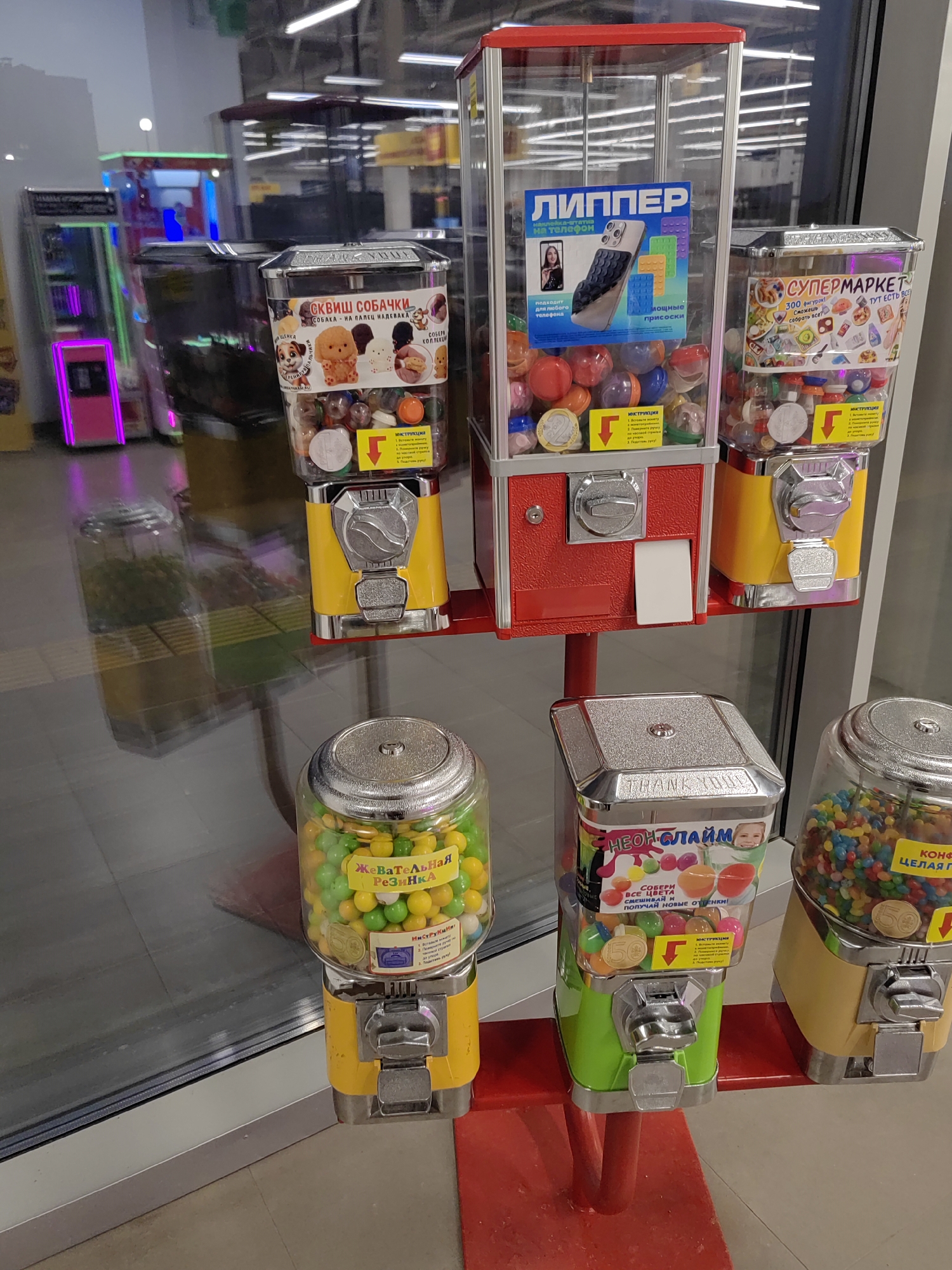 The image shows a display of several gumball or capsule toy machines, likely in a retail environment such as a supermarket or arcade. The machines are arranged in a row and are brightly colored, with clear plastic containers filled with colorful gumballs or capsules. Some machines have labels with Russian text and images advertising their contents. One machine on the far left is yellow and contains yellow and green gumballs. Next to it is a red machine with a clear capsule holder containing red, blue, and yellow capsules. Another machine, also yellow, is to the right and is filled with multicolored gumballs. A green machine in the foreground contains colorful gumballs. In the background, other similar machines and what appear to be arcade games with illuminated screens are visible. The overall environment appears to be indoors, with reflective flooring and overhead lighting.