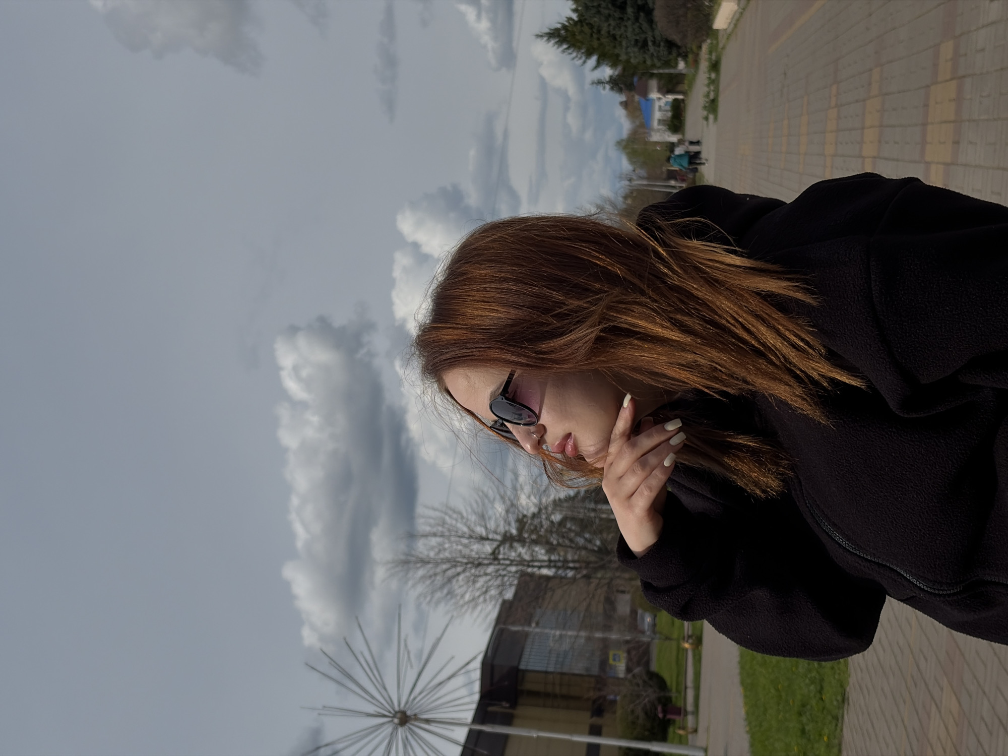 A medium shot captures a person with auburn hair and sunglasses, wearing a black hooded jacket. The person is gazing downward with their hand near their face. The background reveals an urban landscape in Novopokrovskaya, Russia. Buildings, trees, and a modern, dandelion-shaped sculpture are visible. The sky is overcast with gray clouds. Paved walkways suggest a public space like a park or plaza.