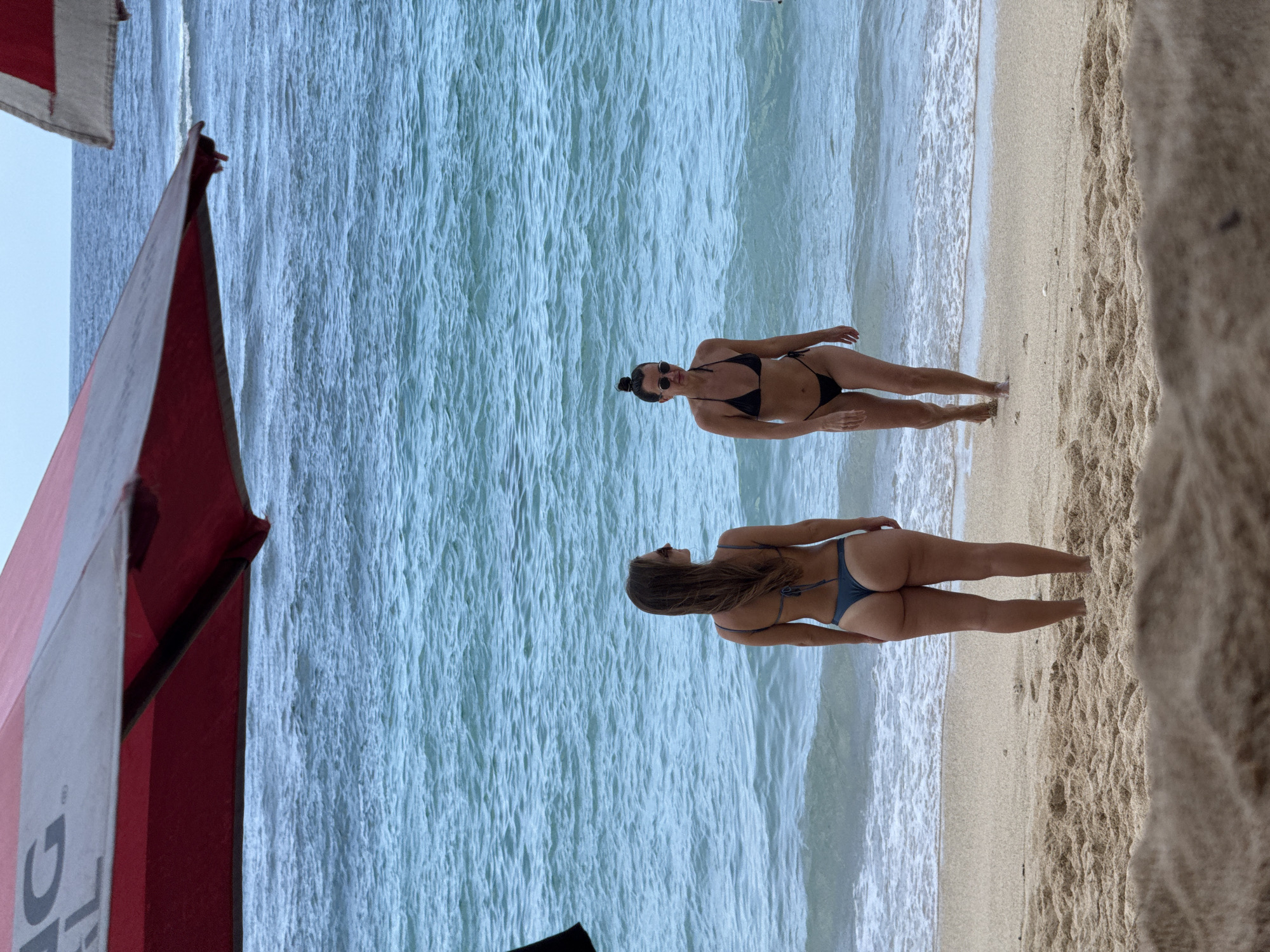The image depicts a beach scene in Kuta Selatan, Indonesia, featuring two women in bikinis. The women are near the shoreline where the ocean meets the sand. One woman wears a black bikini and has her head tilted back. The other woman wears a dark bikini. Parts of a red and white object are visible on the left side of the frame. The sand is light in color, and the ocean water is a pale blue.