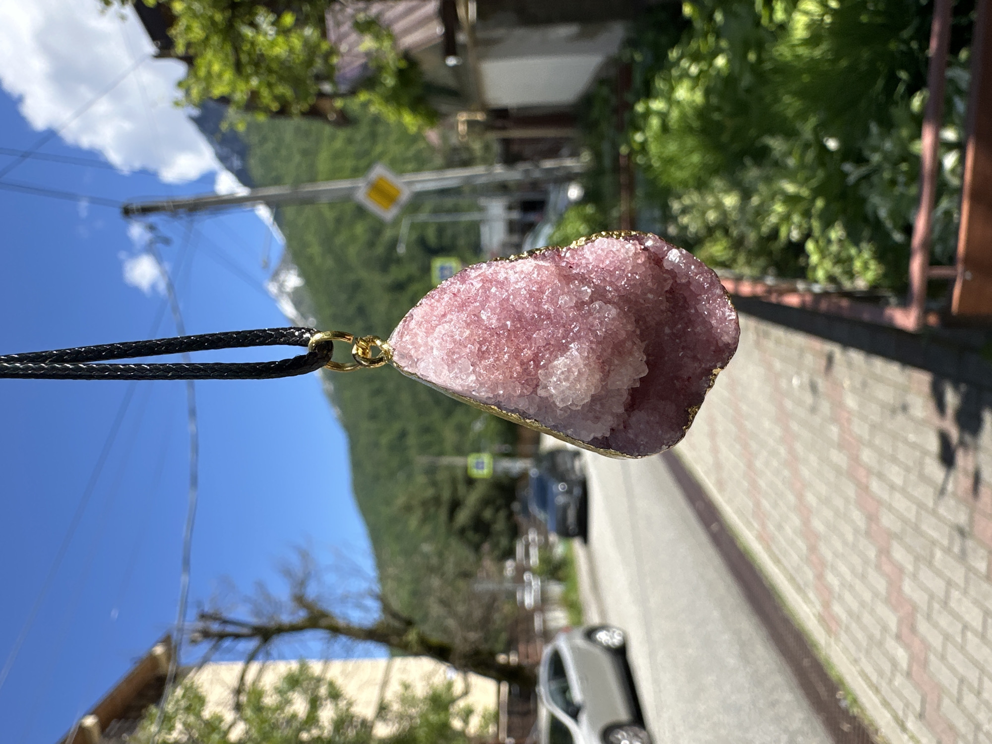 A pink crystalline pendant, intricately detailed with numerous small, sparkling crystals and trimmed with gold-colored metal, is the central subject of this image. It hangs from a black braided cord, sharply focused against a blurred outdoor background.
The setting appears to be a sunny day in a mountainous or hilly region. The sky is bright blue with scattered white clouds, suggesting midday or early afternoon. In the heavily defocused background, lush green mountains rise in the distance, while abundant green trees and foliage frame the middle ground.
A paved road or street is visible, with a light-colored car parked or driving on it. To the right, there's a structure with a brick-patterned facade, possibly a wall or part of a building. A utility pole with overhead wires and a yellow square street sign with an indistinguishable black symbol are also present, indicating an inhabited area or village. The scene combines natural beauty with elements of a developed settlement, consistent with the location context of Krasnaya Polyana, Russia. There are no people visible, nor is any readable text present in the image.