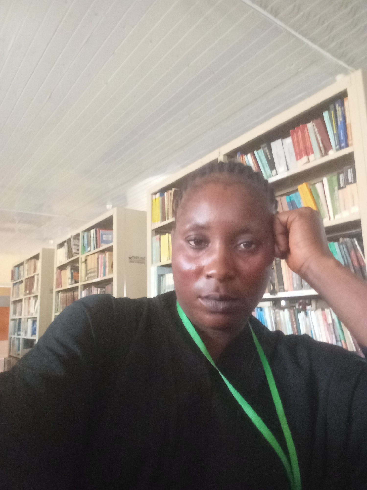 A selfie shot shows a woman with dark skin and braided hair, resting her head on her hand. She wears a black garment and a green lanyard. Behind her, shelves of books indicate she is likely in a library or study. The location may be Akwanga, Nigeria, but there are no specific details in the image confirming this.