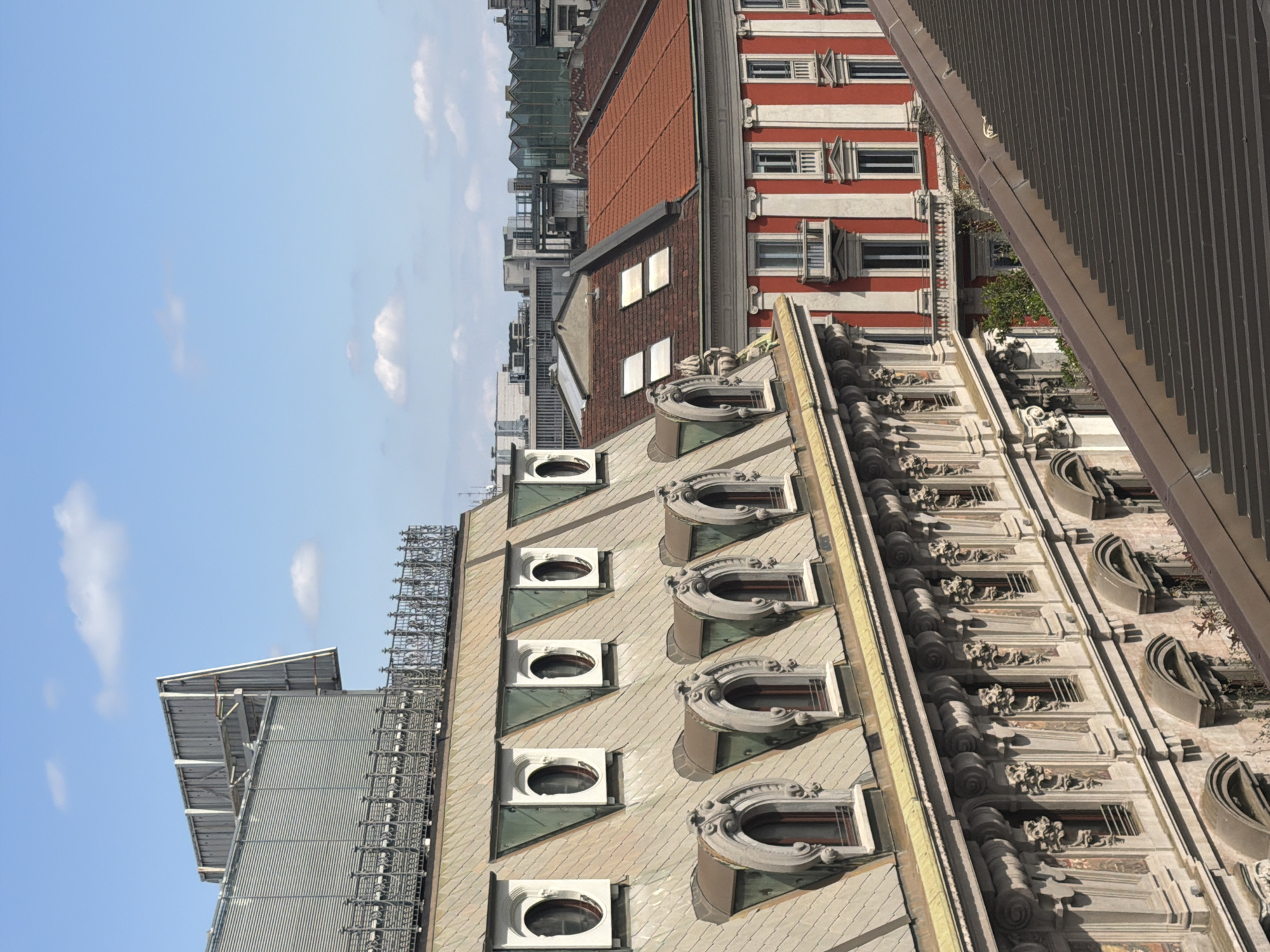 This is an upward-angled view of the architecture of Milan, Italy, including details of rooftop construction and exterior facades against a sky with scattered clouds. On the right is a section of a building with decorative elements. Adjacent to this is a lighter-colored roof with oval-shaped windows extending through. In the background are more buildings of various styles, including one with a red and white striped facade. The sky occupies a significant portion of the frame.