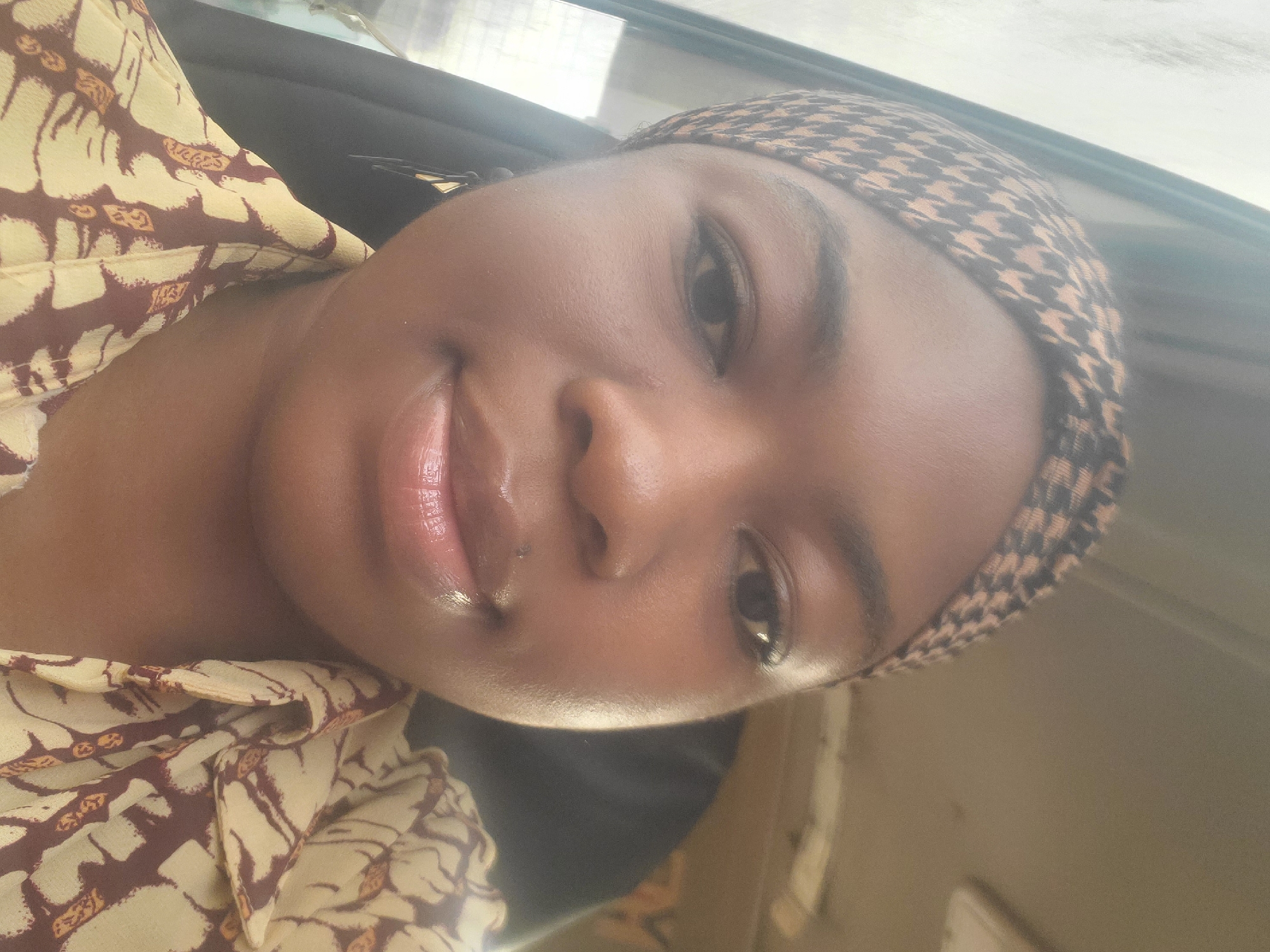 The image is a selfie taken by a young woman in Kakau, Nigeria. She is wearing a headwrap with a houndstooth pattern and a yellow shirt with a brown abstract pattern. Her lips are glossy. The background appears to be the interior of a vehicle, with a window on the left side showing a blurry outdoor scene.