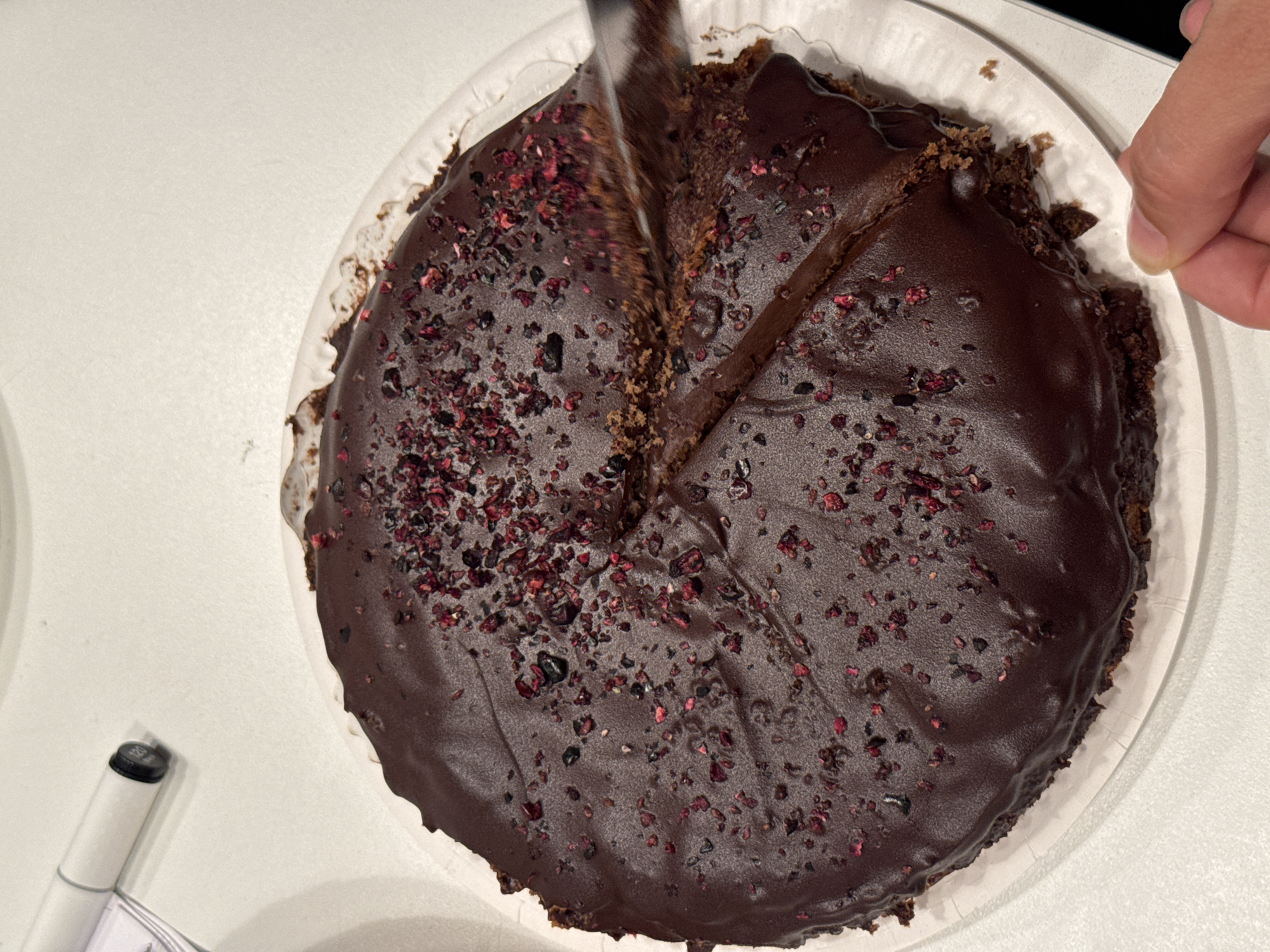 A chocolate cake, topped with freeze-dried raspberry pieces, is being cut with a knife. The cake sits on a white, scalloped-edge paper plate, which is on a white surface. To the upper left of the cake, a white marker pen with a black cap is visible. A person's hand is visible in the lower right corner, holding a slice of the cake as it is being cut. The scene appears to be indoors, possibly a kitchen or dining area. No specific location in Riga, Latvia is depicted.