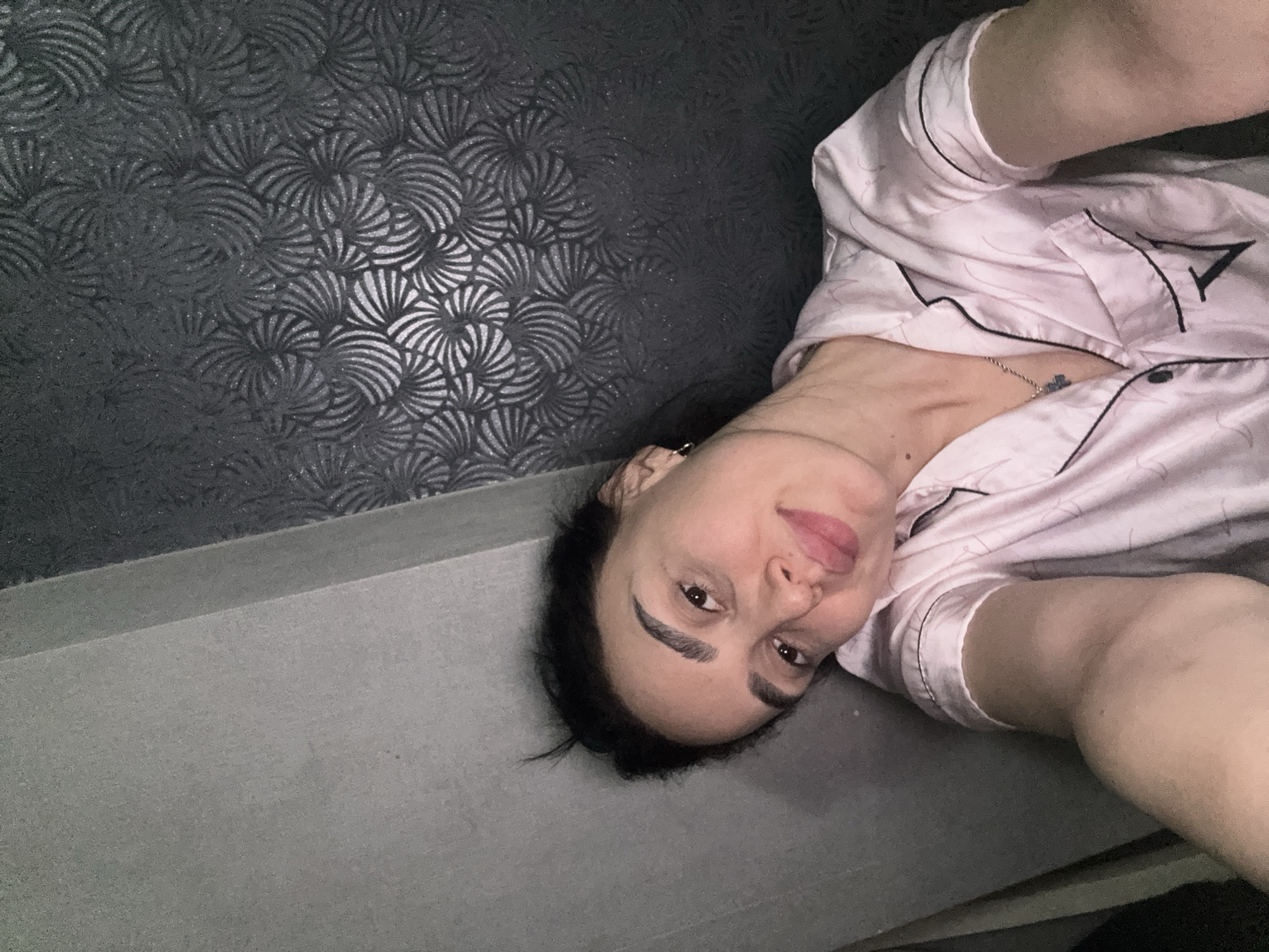 A person is lying on a gray surface next to a wall with a patterned gray covering. The person is wearing light pink pajamas with black piping and a necklace. The location is unknown.