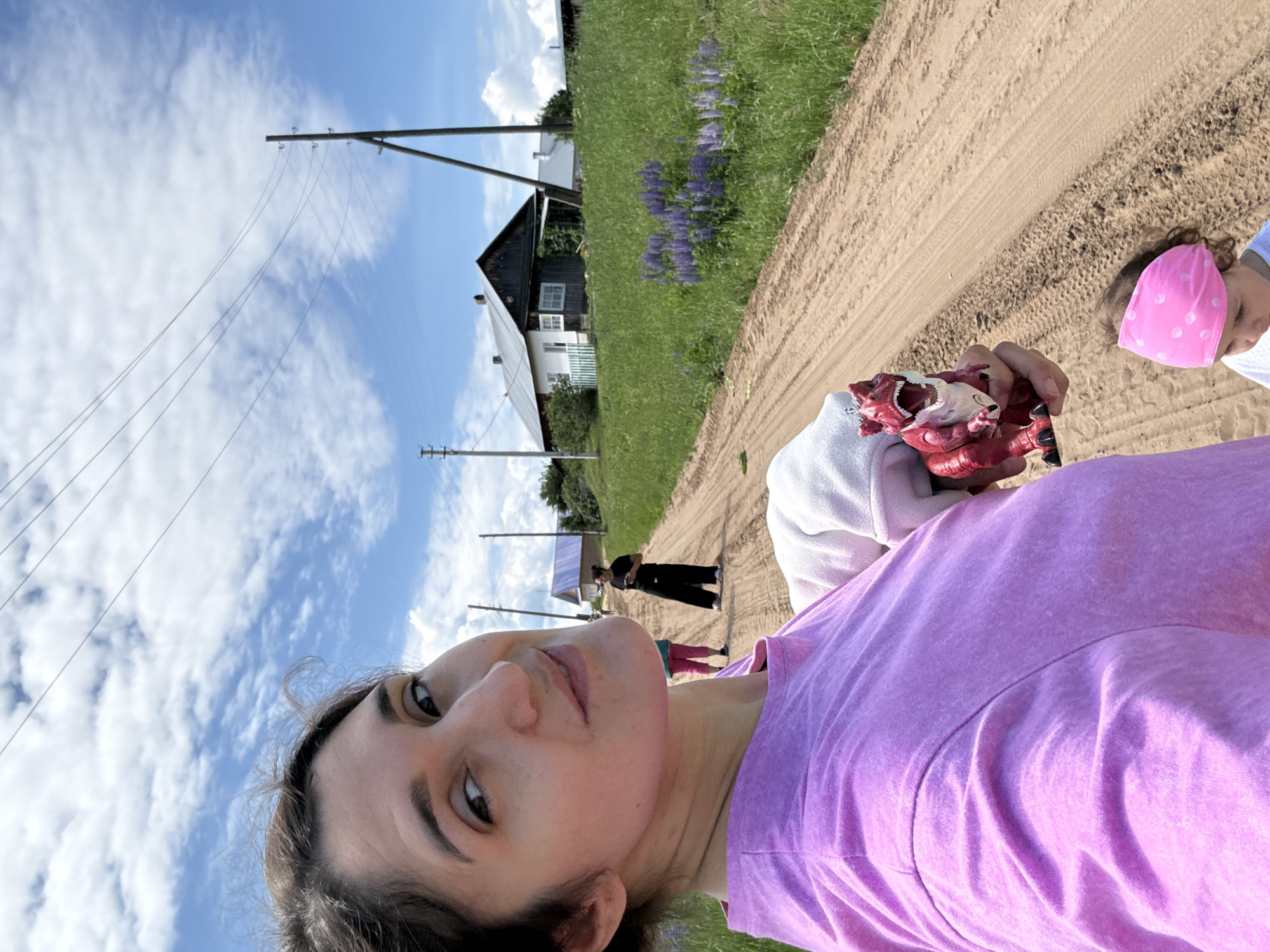 A selfie-style photograph depicts a woman on a dirt road in Уральское, Russia, under a bright, cloudy sky. The woman, appearing to be in her 20s or 30s with dark hair pulled back, occupies the left foreground, looking towards the viewer with a slight smile. She wears a light purple short-sleeved t-shirt. Her left hand holds a red toy Tyrannosaurus Rex with its mouth open, positioned in the lower right foreground. Partially visible below the toy is the head of a young child wearing a pink bandana with white polka dots.
The scene extends along a light brown dirt road with visible tire tracks, receding into the background. To the right of the road, green grass and clusters of purple lupine flowers are present. Further down the road, two individuals are visible: one person in pink pants and a dark top stands on the left side of the road, while another person in black wide-legged pants and a dark top stands further right, appearing to hold a mobile phone.
In the background on the right, two traditional wooden houses are situated. The more prominent house features a white lower facade, dark wooden upper walls, a grey metal roof, and a small white fence with green details. A second house with a lighter roof is partially visible behind it. Several wooden utility poles with overhead power lines traverse the blue sky, which is dotted with scattered white cumulus clouds.