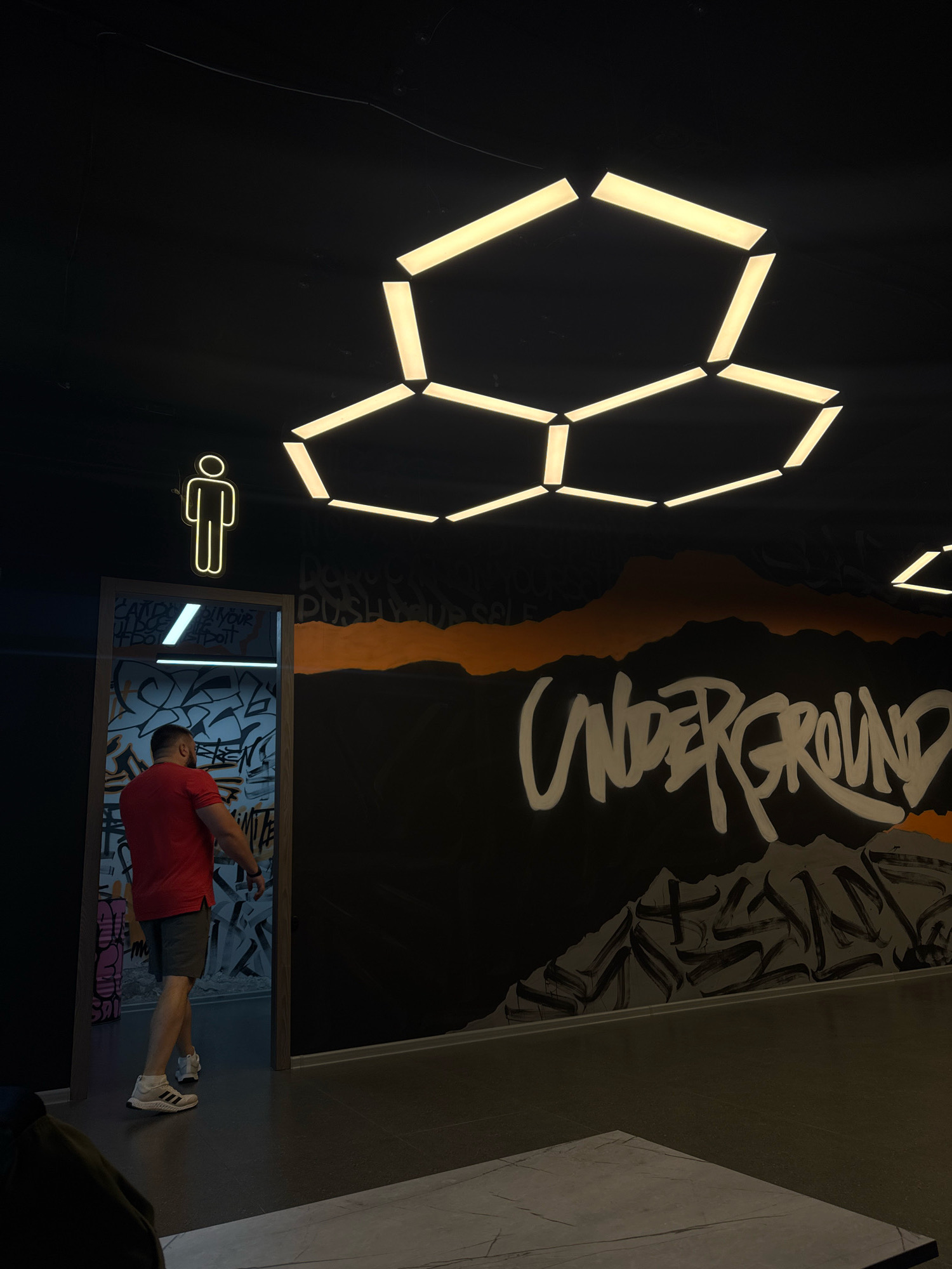 The image depicts the entrance to a restroom, possibly in a commercial establishment in Almaty, Kazakhstan. A man wearing a red t-shirt and grey shorts is walking into the restroom, which is indicated by a neon sign of a male figure above the doorway. The restroom door is open, revealing a space covered in graffiti. The surrounding environment features a dark ceiling with an abstract, hexagonal light fixture. The wall to the right of the restroom entrance is adorned with graffiti, including the word "UNDERGROUND" written in large, stylized lettering, along with mountain-like imagery. The floor appears to be dark tile. The lighting is dim, with the neon sign and the ceiling fixture providing illumination. The overall aesthetic suggests a modern, urban, and possibly edgy venue.