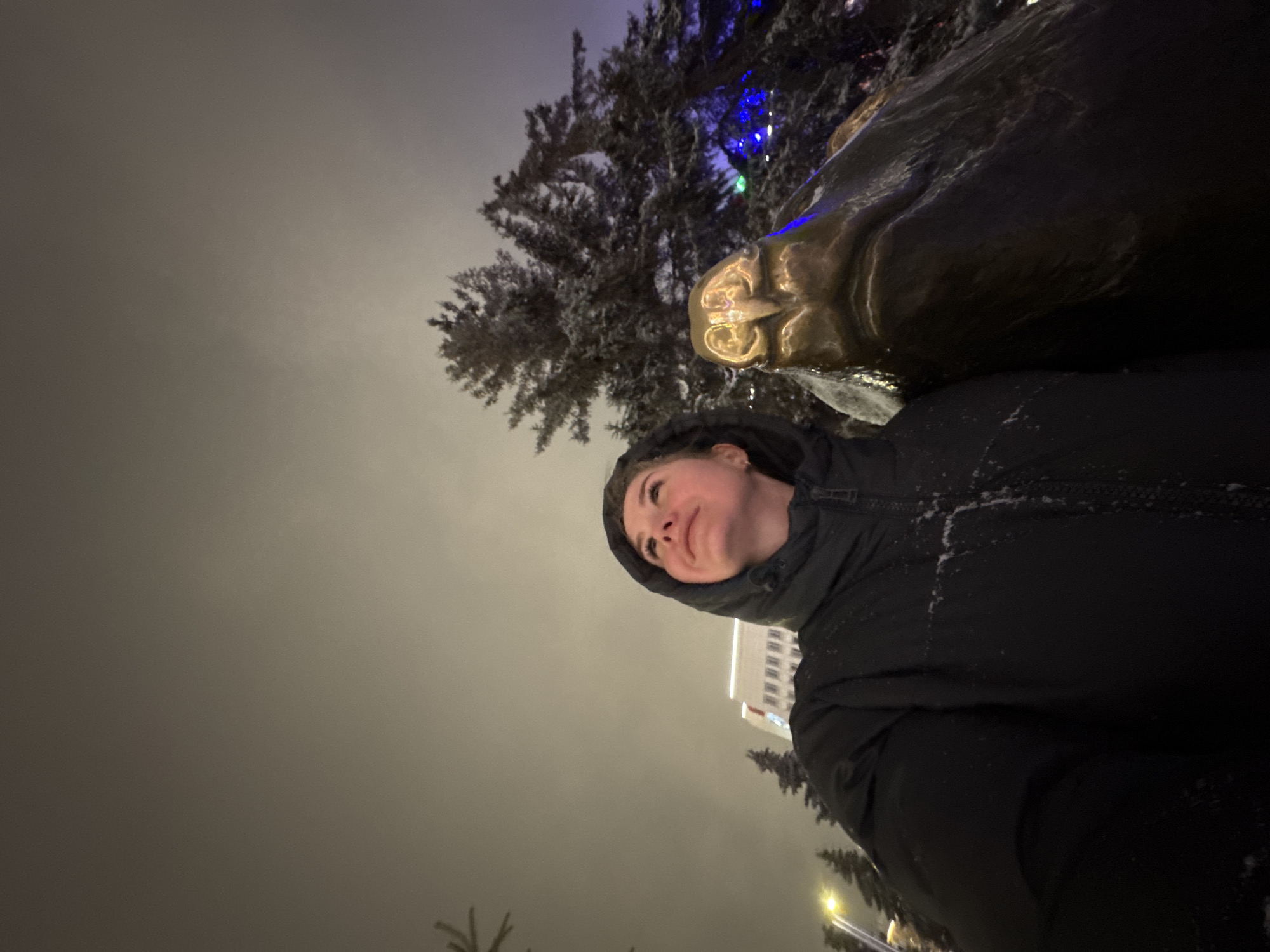 A low-angle shot depicts a woman wearing a black hooded jacket, looking up and slightly to the left at a bronze sculpture of a bear. The sculpture and the woman are in the foreground, partially obscuring a decorated Christmas tree with colored lights behind the sculpture. In the background, partially visible, is a light-colored building with multiple windows, likely a government or municipal building. The scene is located in the city of Perm, Russia, specifically at the bear sculpture which is a known landmark. The environment appears to be nighttime or dusk with an overcast sky. There is snow visible on the sculpture and possibly on the woman's jacket, indicating a cold climate. The perspective is such that the woman is close to the sculpture, creating an intimate feeling.