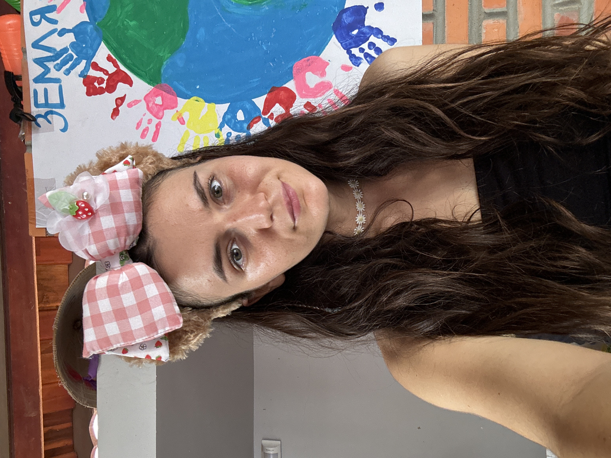 A selfie of a woman with long, dark brown hair, wearing a black tank top, a daisy choker, and a novelty headband with ears and a large pink and white gingham bow with a strawberry embellishment. She is indoors, standing in front of a painted mural featuring handprints and a depiction of Earth. A sign with the word "Земля" is partially visible above the mural. The location appears to be Mũi Né, Vietnam.