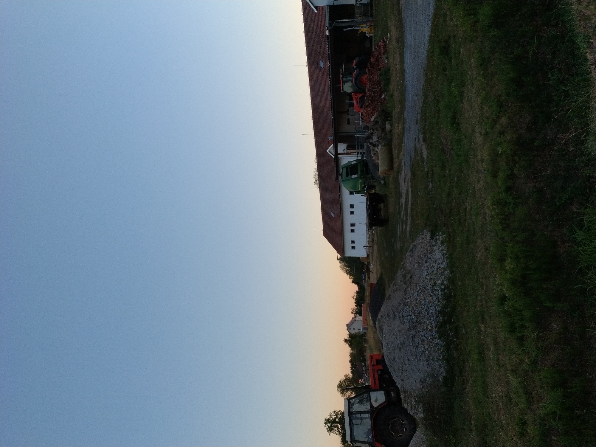The image captures a rural scene at dusk in Dobev, Czechia. In the foreground, a large pile of gravel sits on a grassy area. To the left, a red and white tractor is parked. Behind the gravel pile, a long, white building with a red tiled roof dominates the midground, appearing to be a barn or agricultural structure. A green baler is positioned in front of this building, and to its right, another red tractor is visible. Several piles of bricks and lumber are scattered in front of the building. Trees line the horizon, and in the distance, a few houses can be seen, one of which is white with a light-colored roof. The sky above is a clear gradient of blue, transitioning to a soft orange hue near the horizon. The overall impression is that of a working farm during twilight.