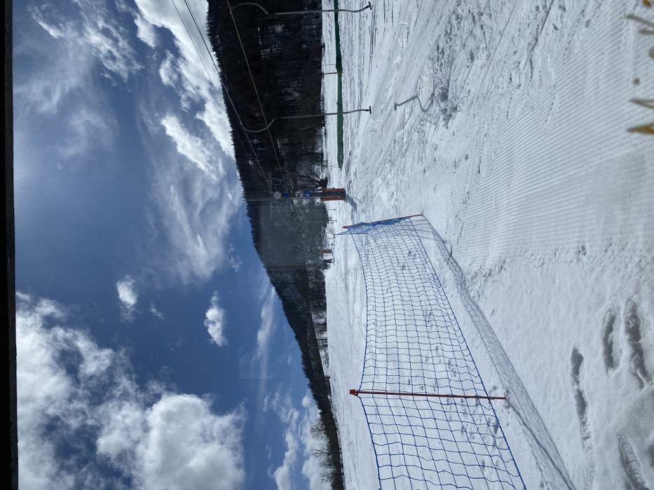 The image depicts a snow-covered ski slope in La Salle-les-Alpes, France, under a partially cloudy blue sky. A blue plastic safety net with red support poles extends across the lower left and mid-ground, casting distinct shadows on the snow. The snow in the foreground shows parallel grooming patterns, while areas near the net display footprints. In the mid-distance, several indistinct figures are visible on the slope, appearing to move towards the base of a ski lift. The ski lift system includes dark pylons and cables ascending the mountain, with some empty lift chairs or T-bars hanging. Orange safety markers are also present near the lift's base. The background consists of a snow-dusted mountain slope densely forested with dark trees. The scene is illuminated by bright sunlight, and the image exhibits reflections or smudges consistent with being photographed through a window or clear barrier, with a dark bar at the top and a faint yellow-orange blur at the bottom right.