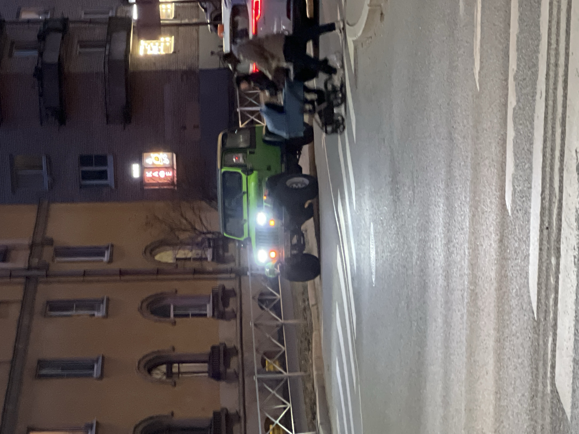 The image captures a nighttime urban street scene in Saint Petersburg, Russia. A bright green, lifted Jeep Wrangler-style vehicle with its headlights illuminated is positioned on a paved road, facing left, near a marked pedestrian crossing. To its right and slightly behind, a white SUV with its red taillights illuminated is partially visible. Two individuals are actively crossing the street from right to left on the zebra crossing. The person on the left is pushing a light blue, four-wheeled baby stroller and is dressed in dark clothing. The second person, walking to the right of the stroller, wears a light-colored jacket, dark pants, and white shoes. A metal barrier fence with a recurring 'X' pattern separates the road from the sidewalk area on the left. In the background, two multi-story buildings are prominent. The building on the left features a yellow-tan facade with multiple arched and rectangular windows, some of which are illuminated. The building on the right is a darker, brick-faced structure with visible balconies. On a lower floor of this brick building, a vertical neon sign displays "КАФЕ" in red Cyrillic letters, accompanied by a smaller illuminated symbol, indicating a commercial establishment. The scene is lit by vehicle headlights, taillights, and interior lights from the buildings, creating artificial illumination against the dark environment.